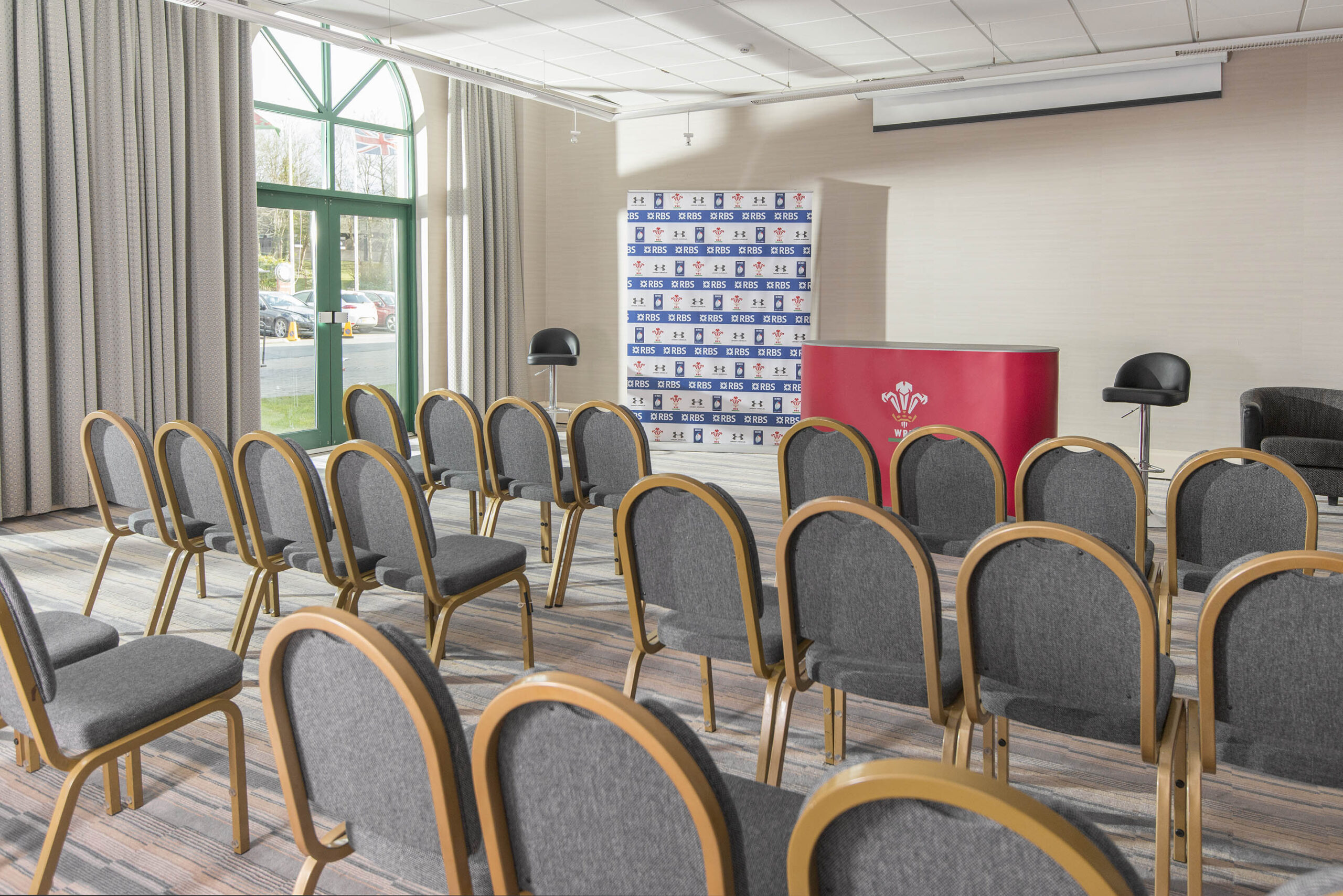 Empty conference room with rows of gray chairs facing a podium and backdrop with logos. Large window and curtains on the left.