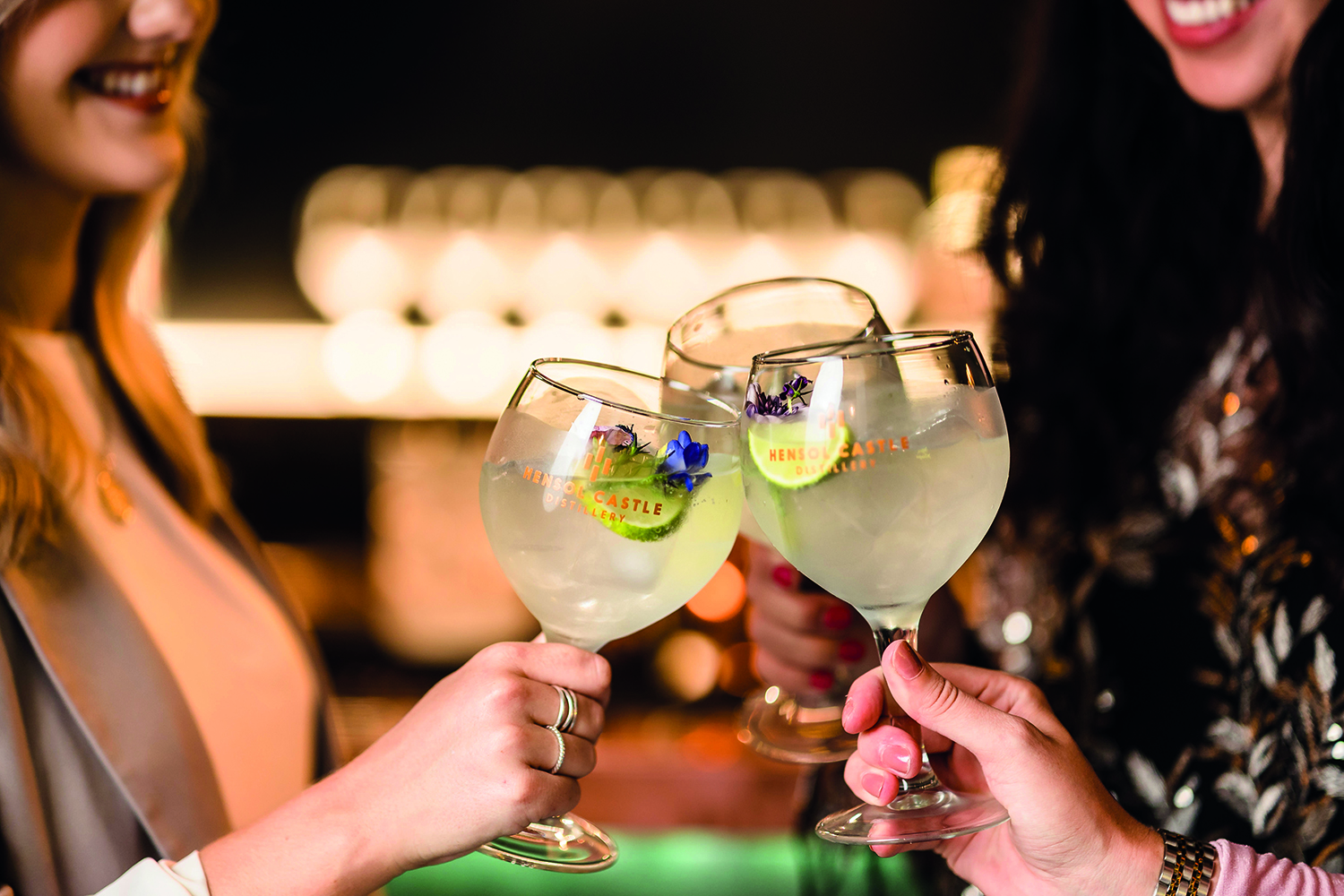 Three people clinking large glasses filled with a light green drink, garnished with lime slices and purple flowers.