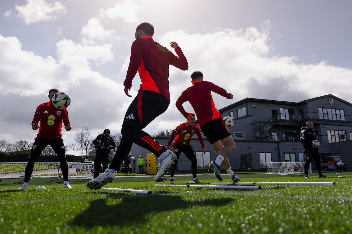 Soccer players in red shirts practice on a field with agility poles and balls under a partly cloudy sky near a large building.