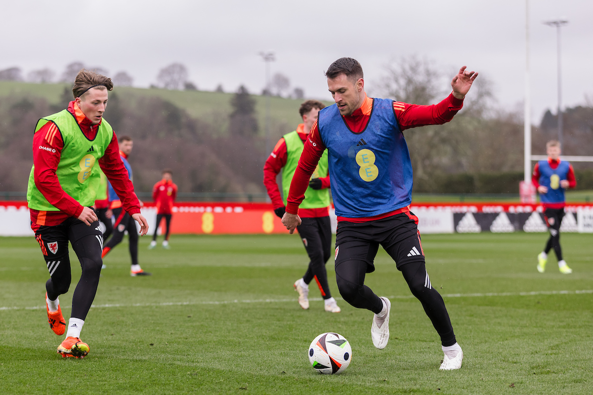 Soccer players in colorful vests practice on a grassy field, with visible hills and trees in the background.