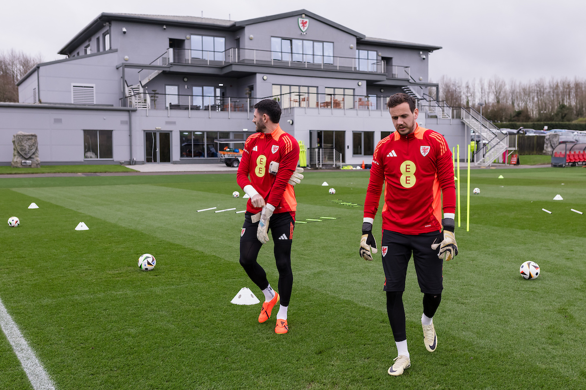 Two soccer players in red jerseys walk on a grass field in front of a modern training facility, with scattered cones and balls around.