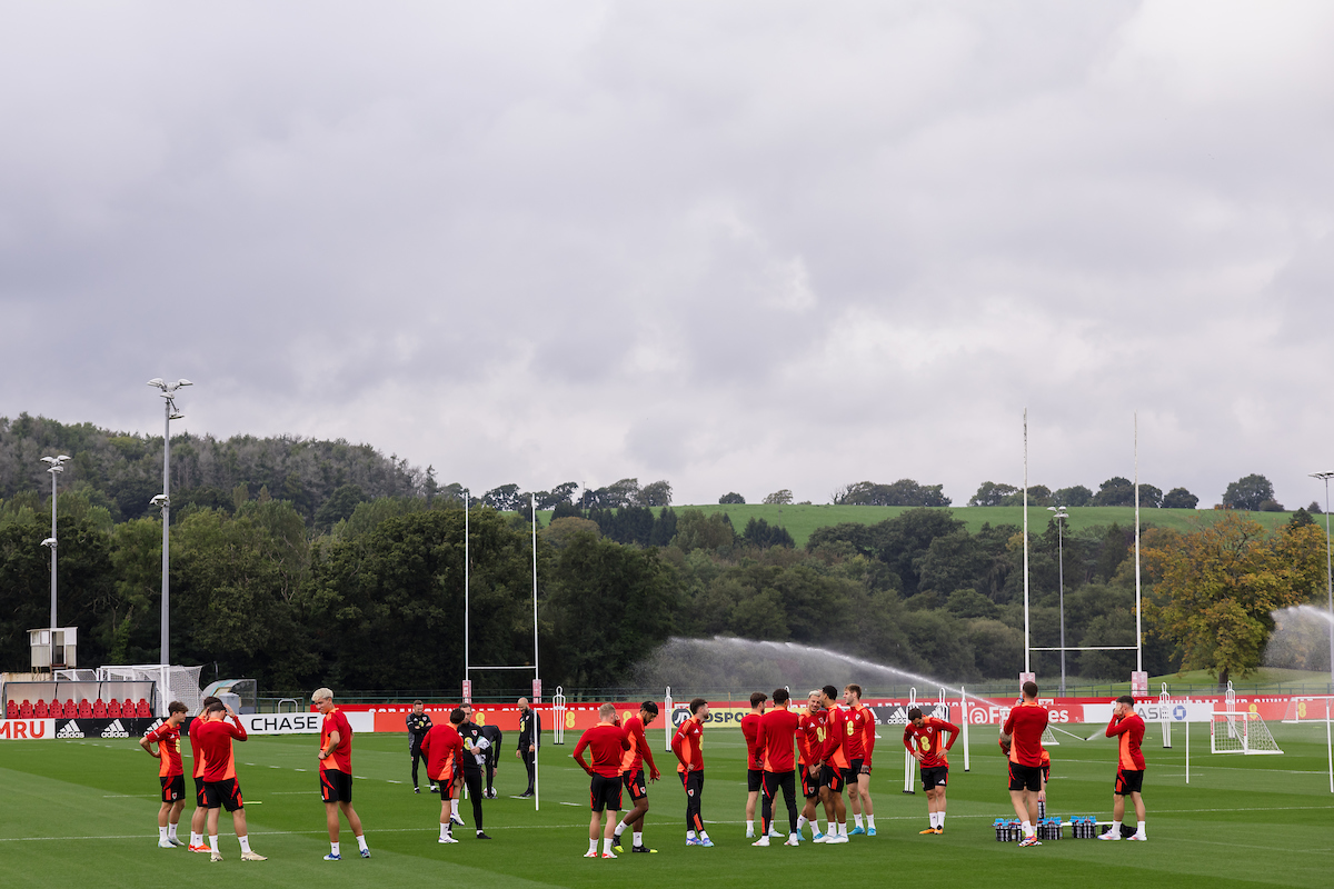 Soccer team in red jerseys gathered on a field, with sprinklers active in the background, surrounded by trees and a cloudy sky.