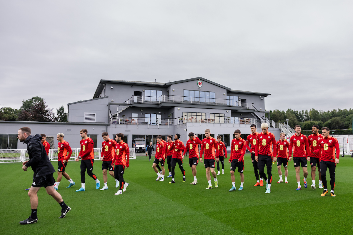 A group of soccer players in red uniforms walk on a green field near a modern gray building under a cloudy sky.