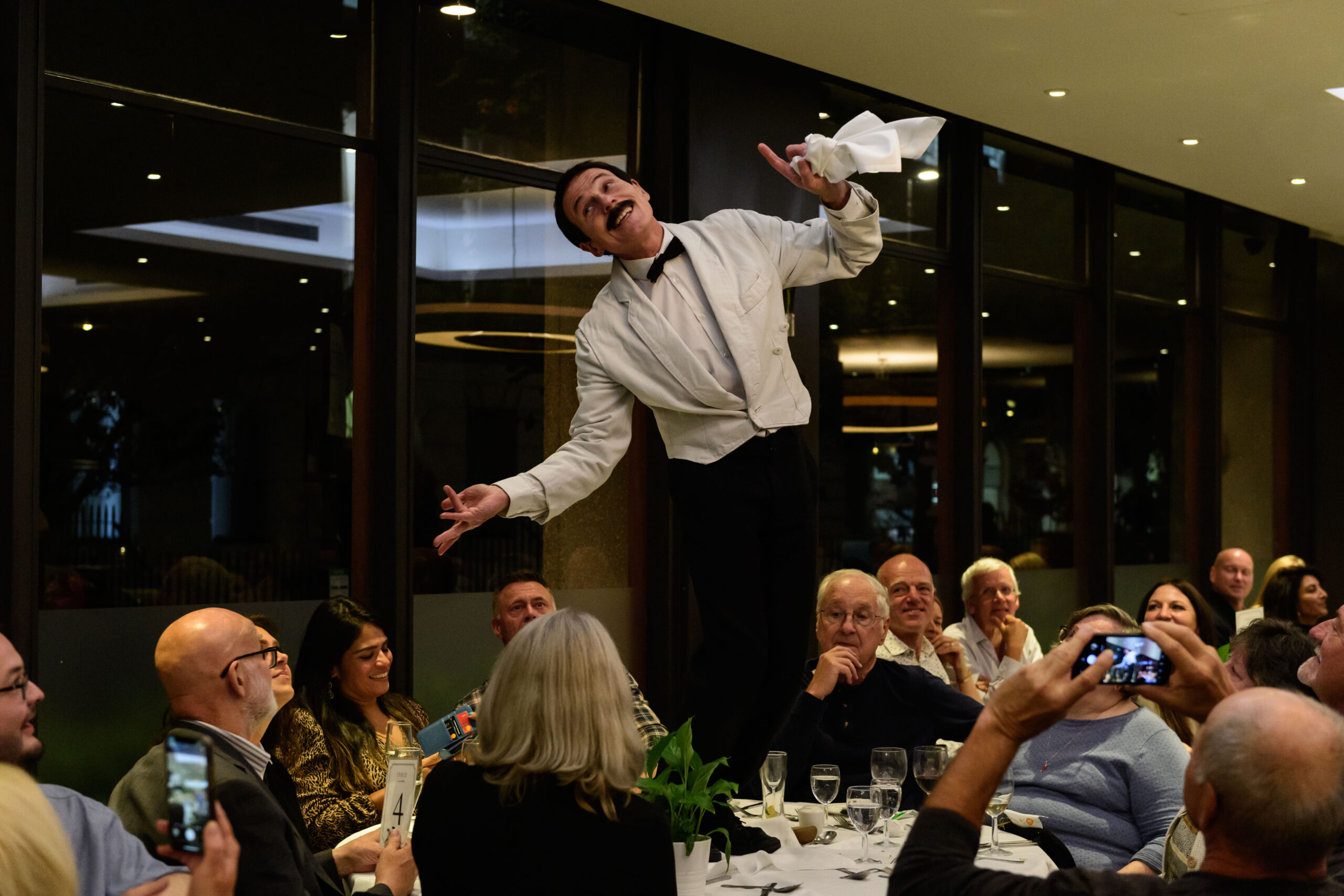 Performer dressed as a waiter entertains a group of diners by standing on a table. Several people are watching, some taking photos with their phones.