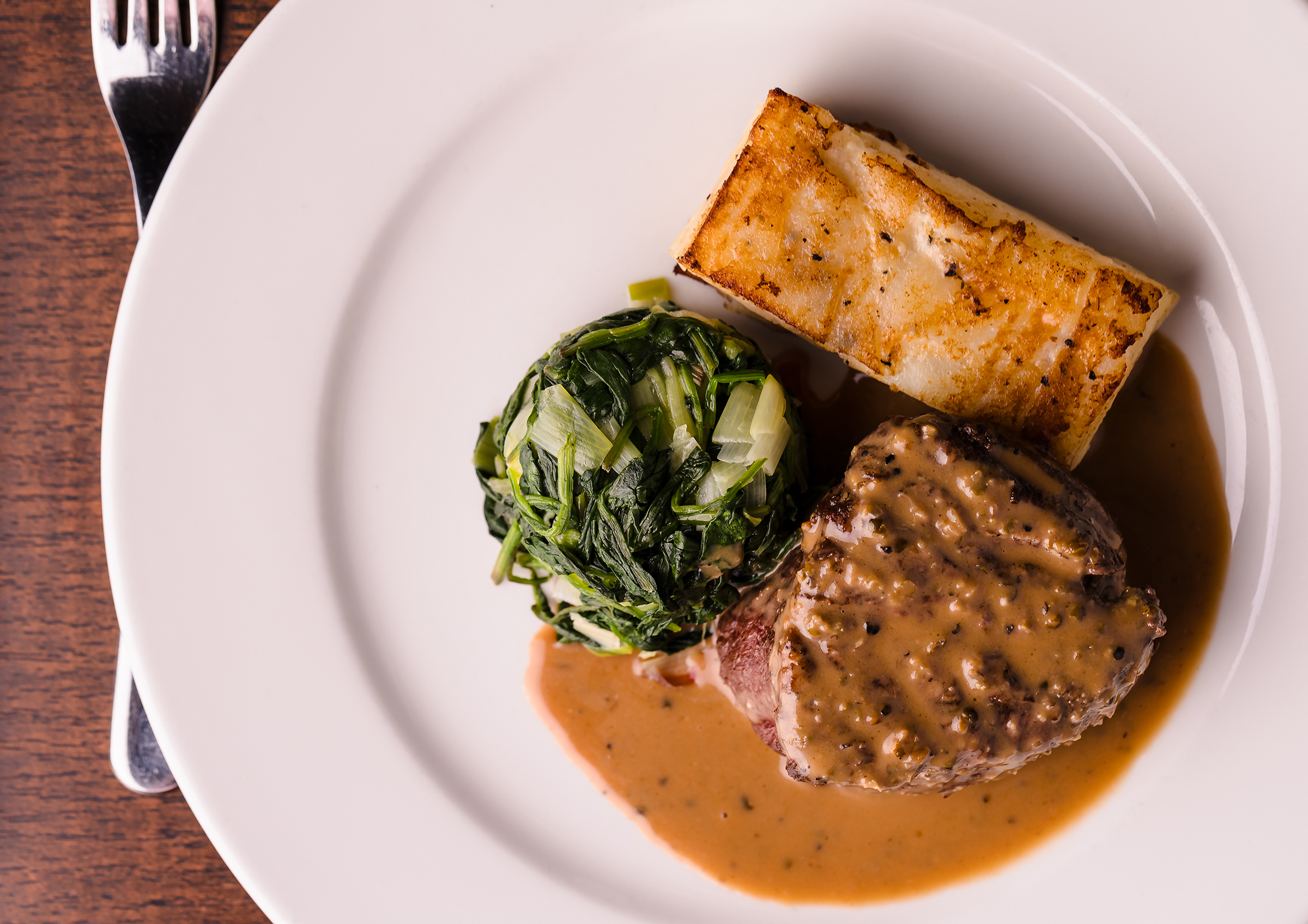 A plate with steak in peppercorn sauce, a portion of sautéed greens, and a slice of potato gratin, on a wooden table with a fork.