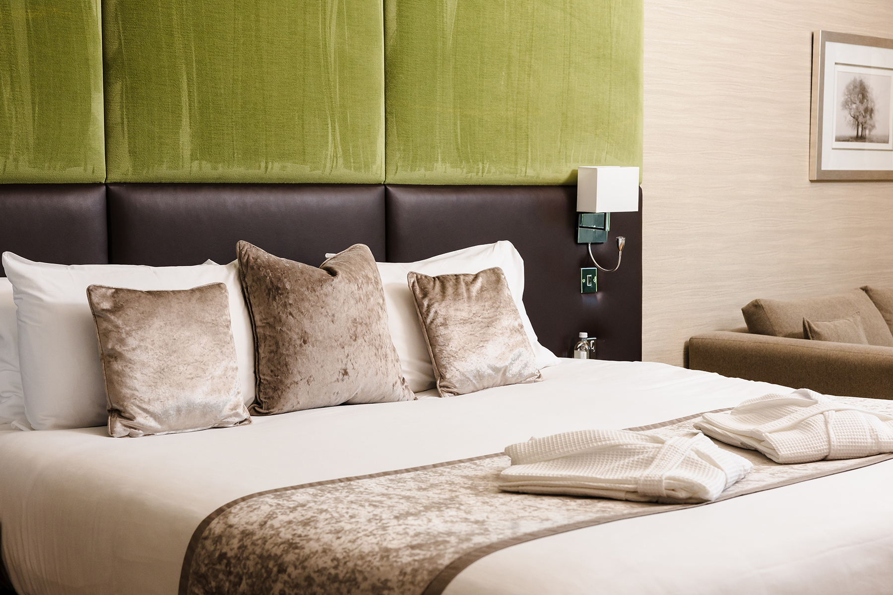 A hotel room features a neatly made bed with green and brown accents, including pillows and a throw. A folded robe rests on the bed. A framed landscape image is on the light-colored wall.