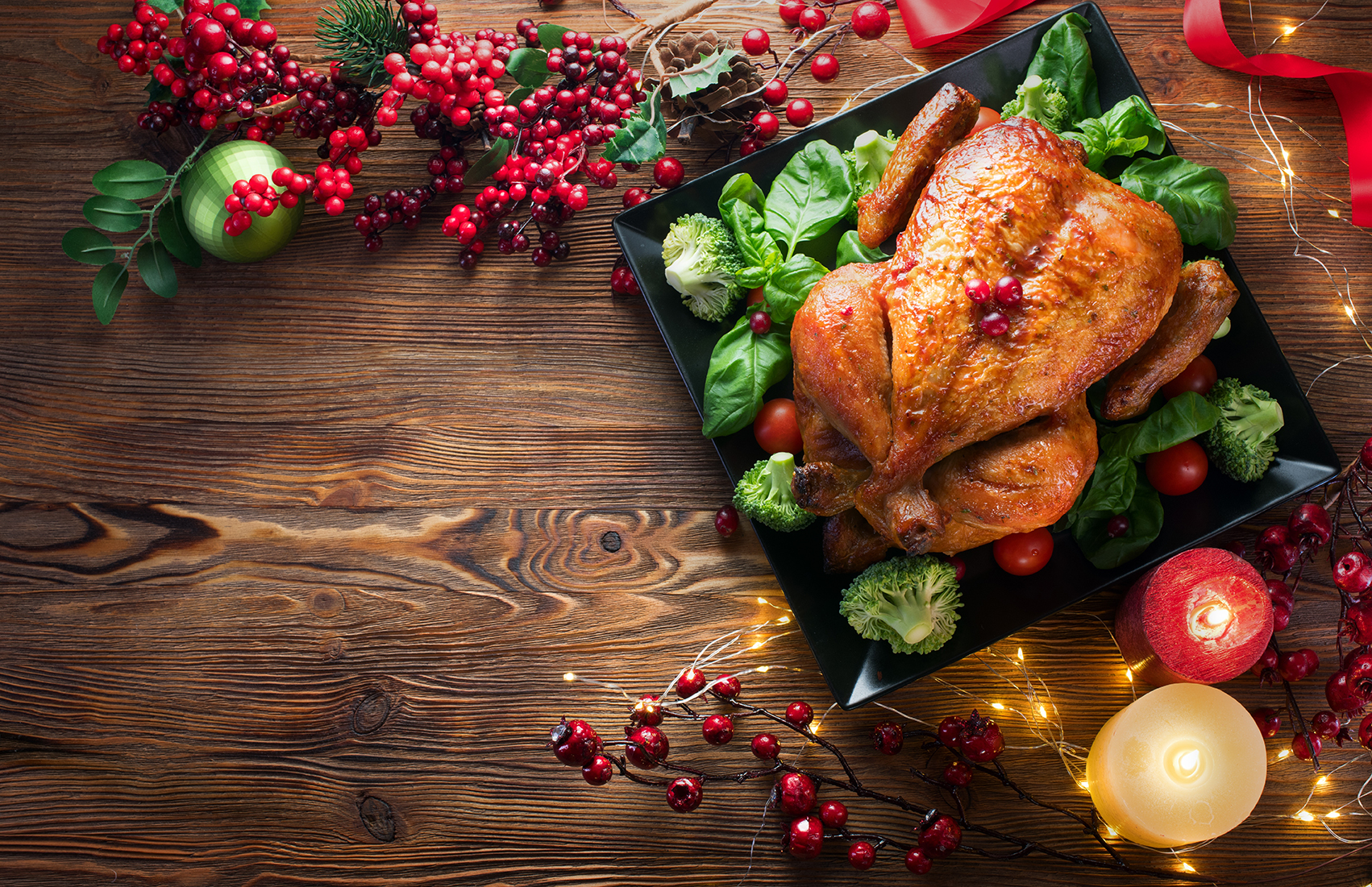 A roasted chicken on a black platter surrounded by greens and tomatoes. The table is decorated with red berries, a candle, and Christmas ornaments.