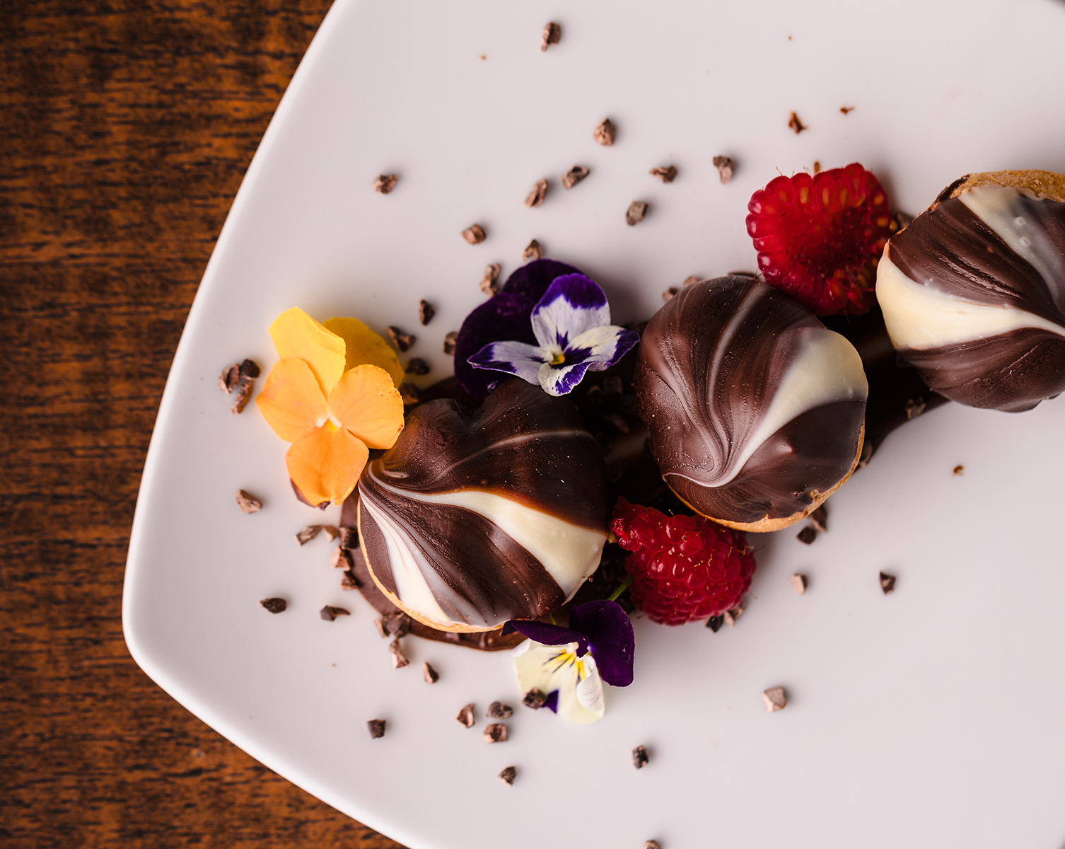 Chocolate-covered dessert balls with white swirls, garnished with edible flowers, raspberries, and cocoa nibs on a white plate.