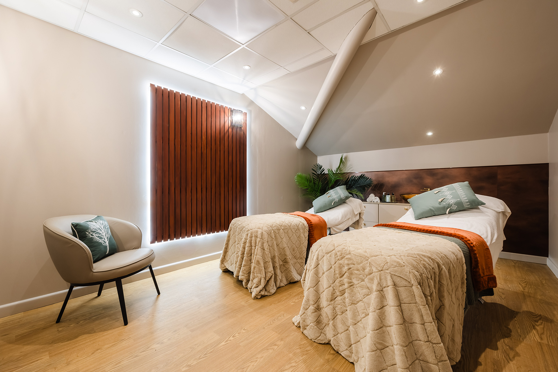 A spa room with two massage beds, soft beige blankets, and green cushions. A chair with a cushion is on the left, and a potted plant is in the background.