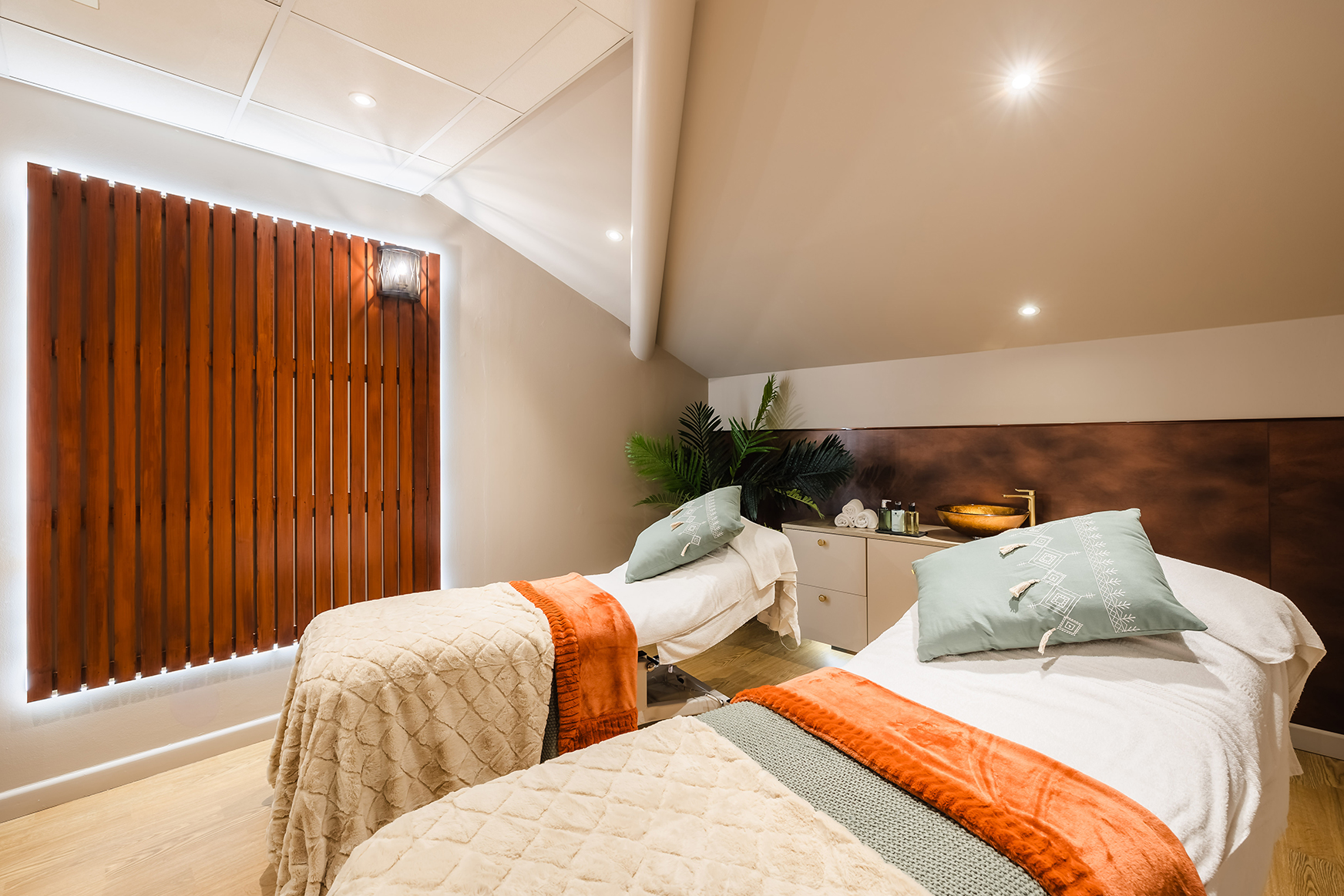 Two massage beds with pillows and blankets in a softly lit room, featuring a wooden wall accent and potted plants.