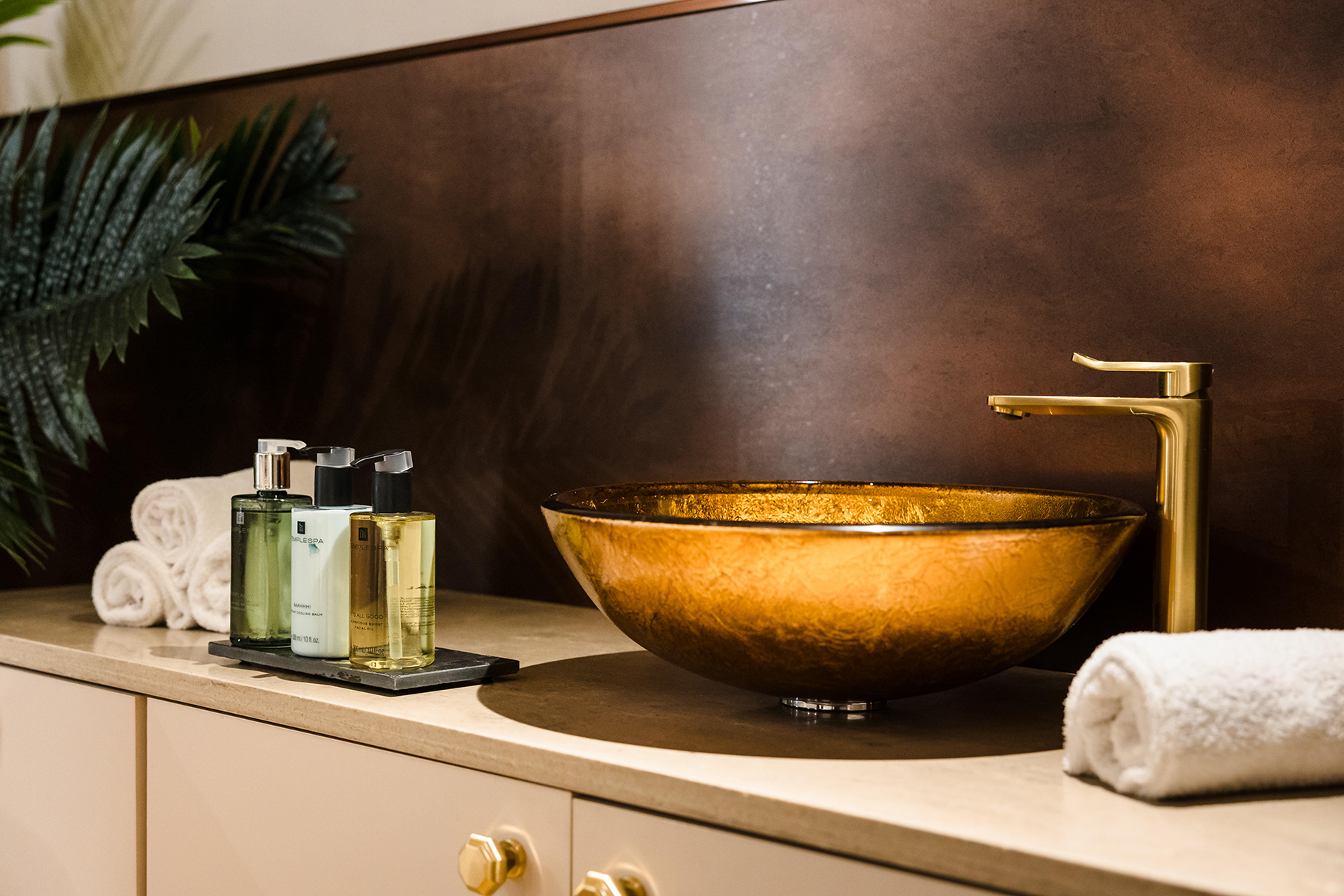 A bathroom with a gold washbasin, sleek faucet, and a set of toiletries next to rolled white towels on a beige countertop.