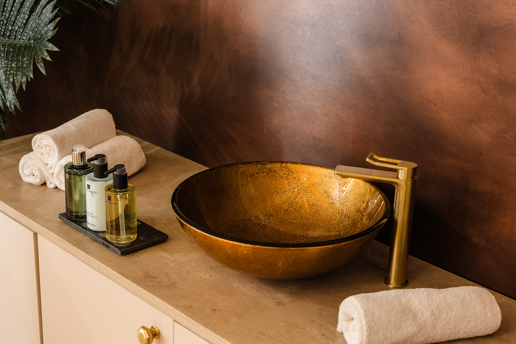 A bathroom sink setup with a round gold basin, gold faucet, rolled white towels, and a tray with toiletry bottles on a beige countertop against a brown textured wall.