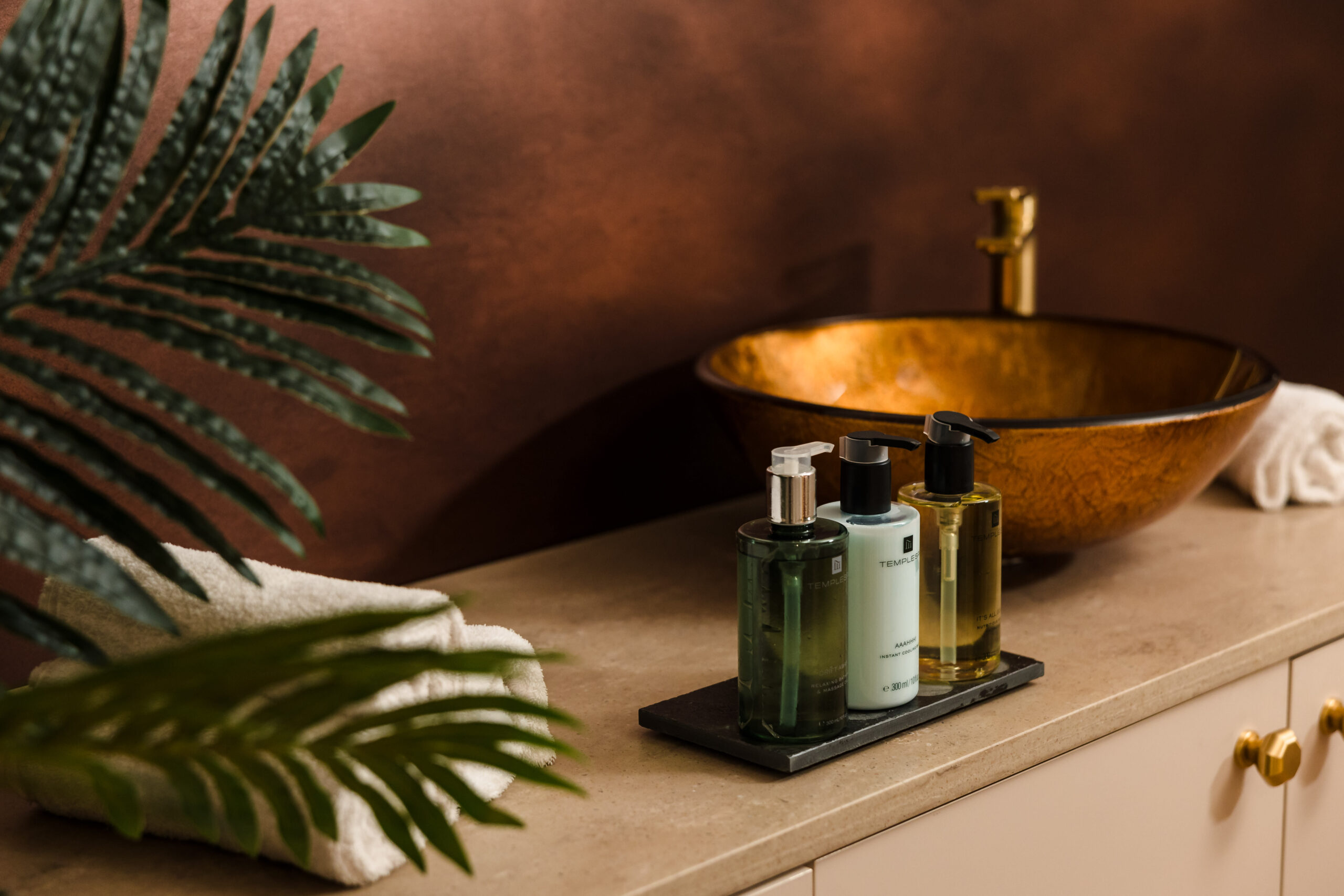 Bathroom sink with three soap dispensers on the counter, a brown basin with a gold faucet, a white towel, and green plant decoration.