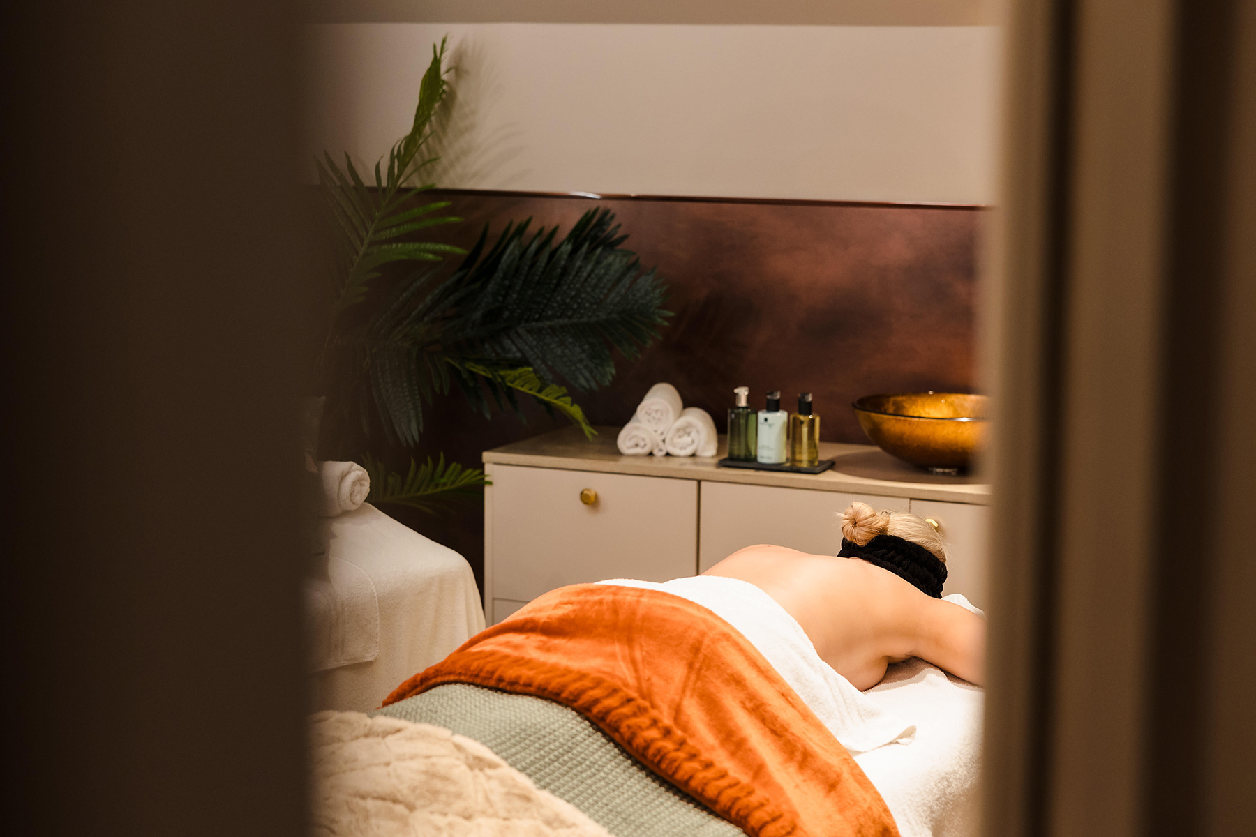 A person lies face down on a massage table, covered with a towel. Nearby, there are rolled towels, bottles, and a gold bowl on a cabinet, with a plant in the background.