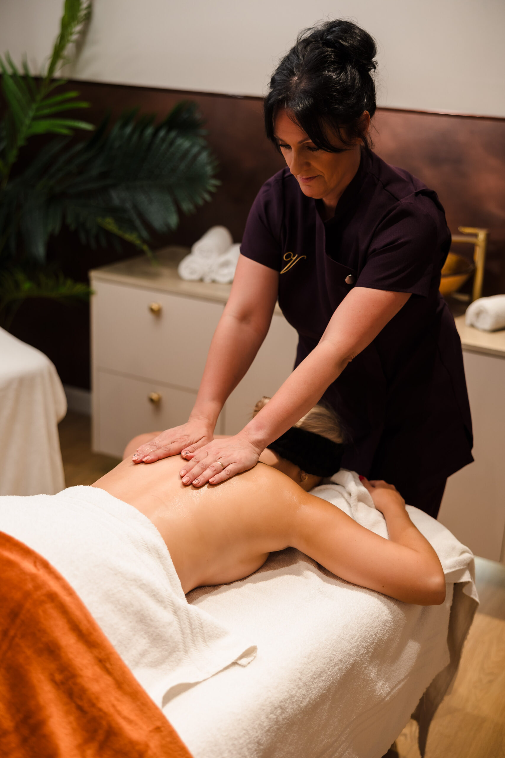 A massage therapist in uniform is giving a back massage to a person lying face down on a massage table, covered with a white towel in a spa setting.