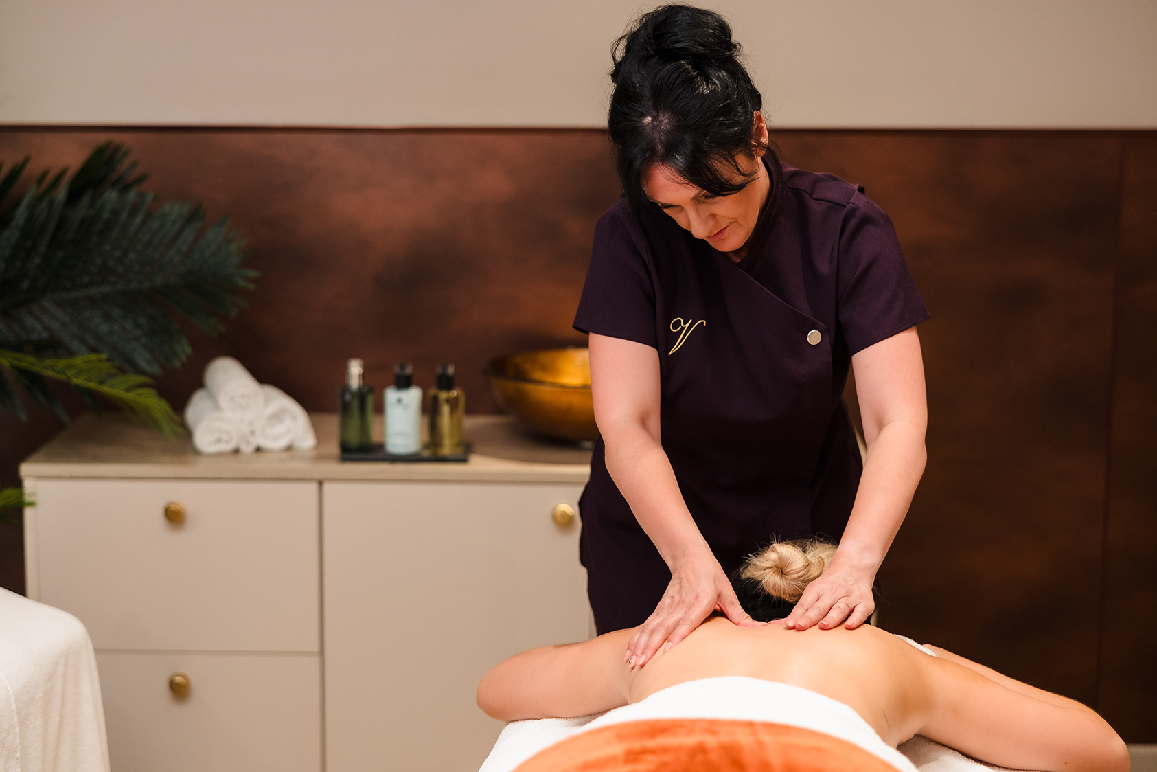 A massage therapist gives a back massage to a client lying face down on a massage table. The room includes soothing decor, towels, and massage oils.