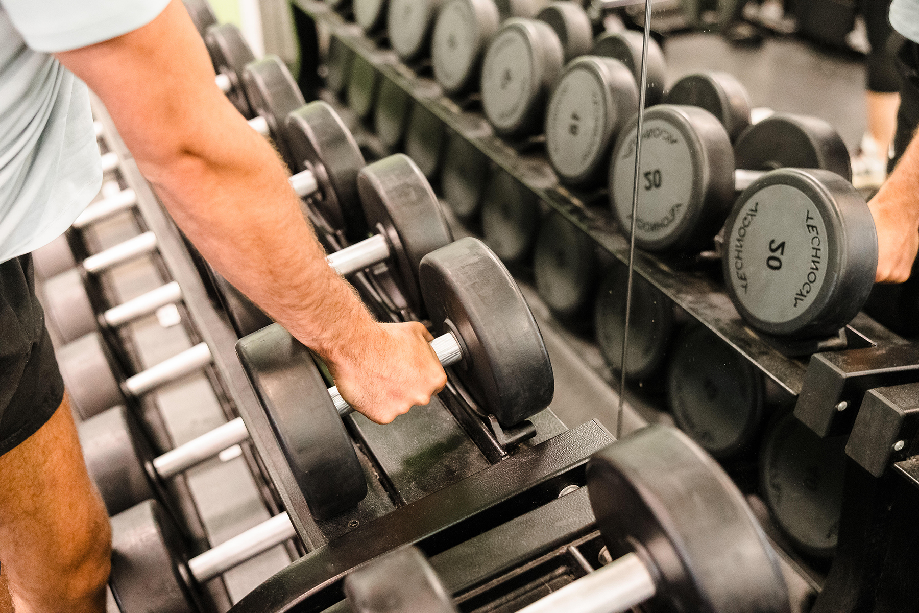 A person grabs a dumbbell from a rack in a gym, with a mirror reflecting the weights.