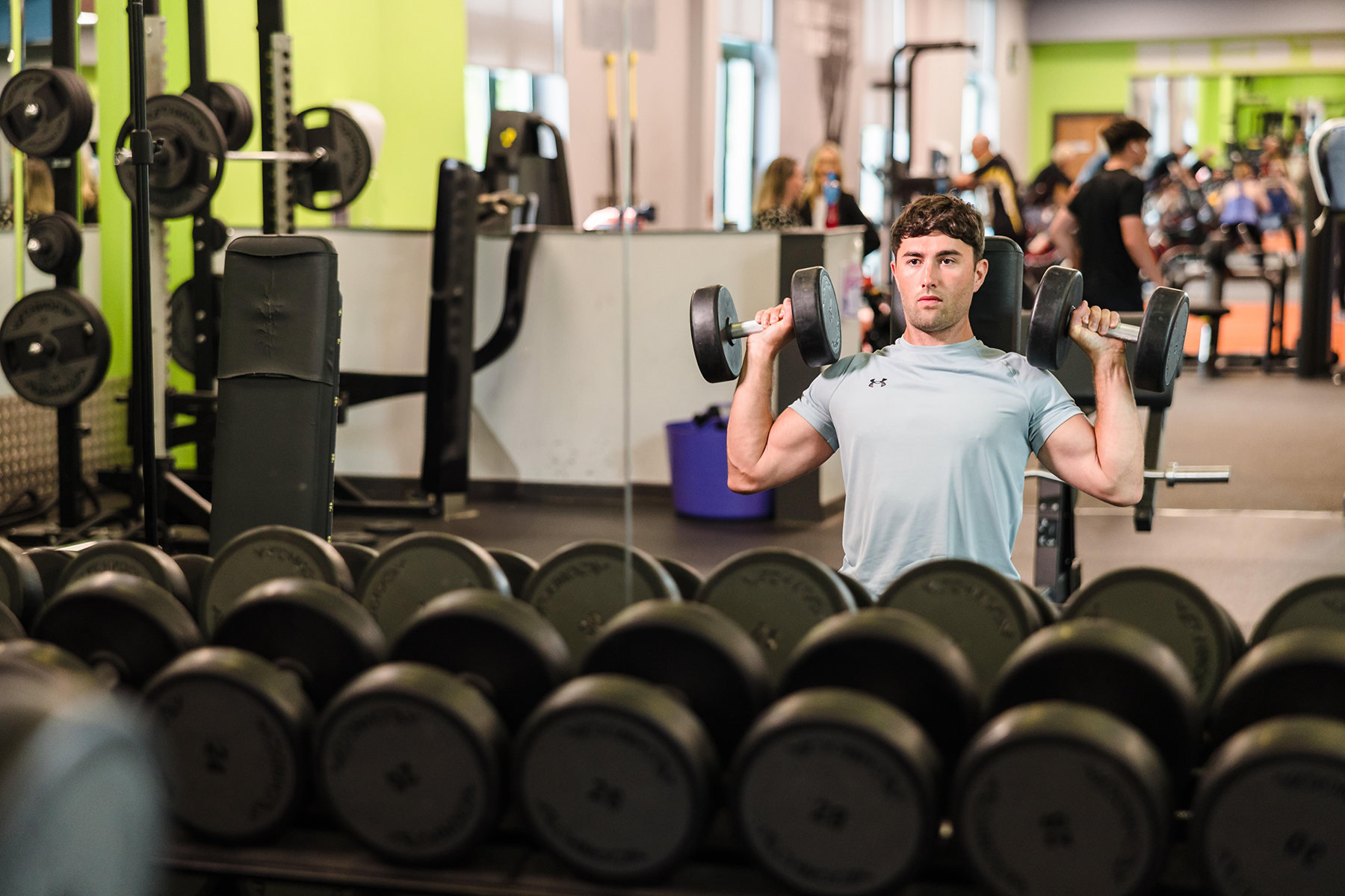 A person in a gym performs a shoulder press exercise with dumbbells in front of a mirror. Rows of weights are visible in the foreground.