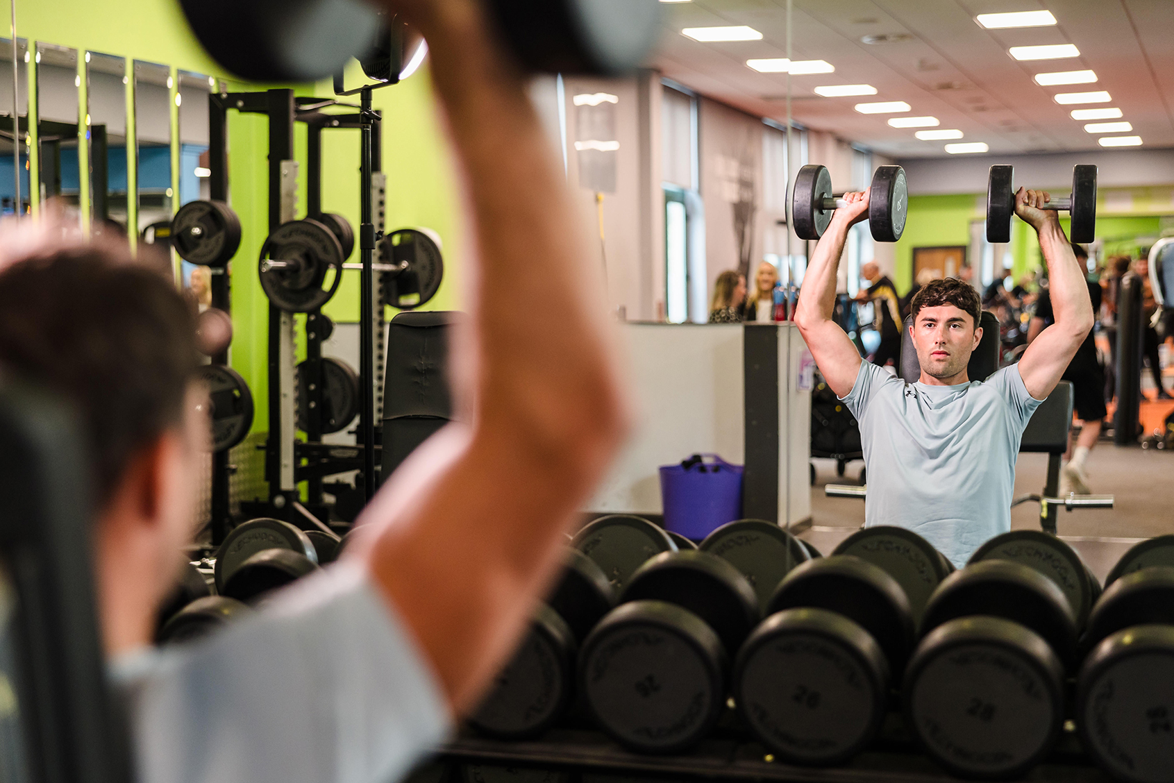 A person in a gym lifts dumbbells overhead while looking at their reflection in a mirror, surrounded by various exercise equipment.