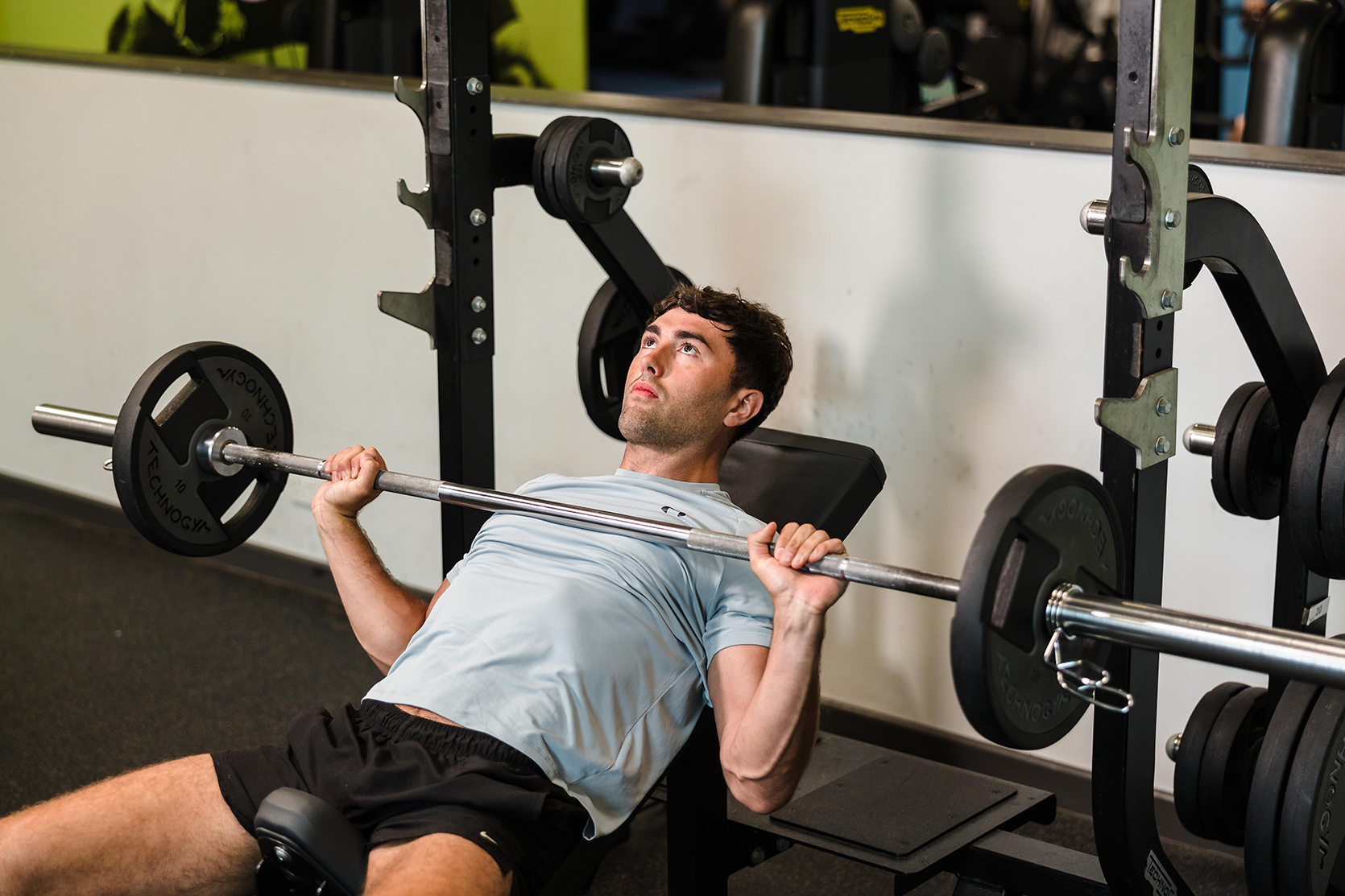 Person in a gym on an incline bench, lifting a barbell with weights.