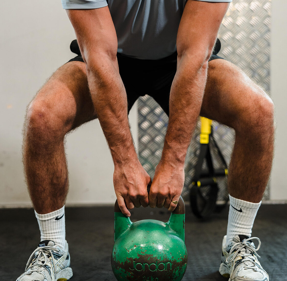 Person in athletic wear lifting a green kettlebell while squatting in a gym setting.