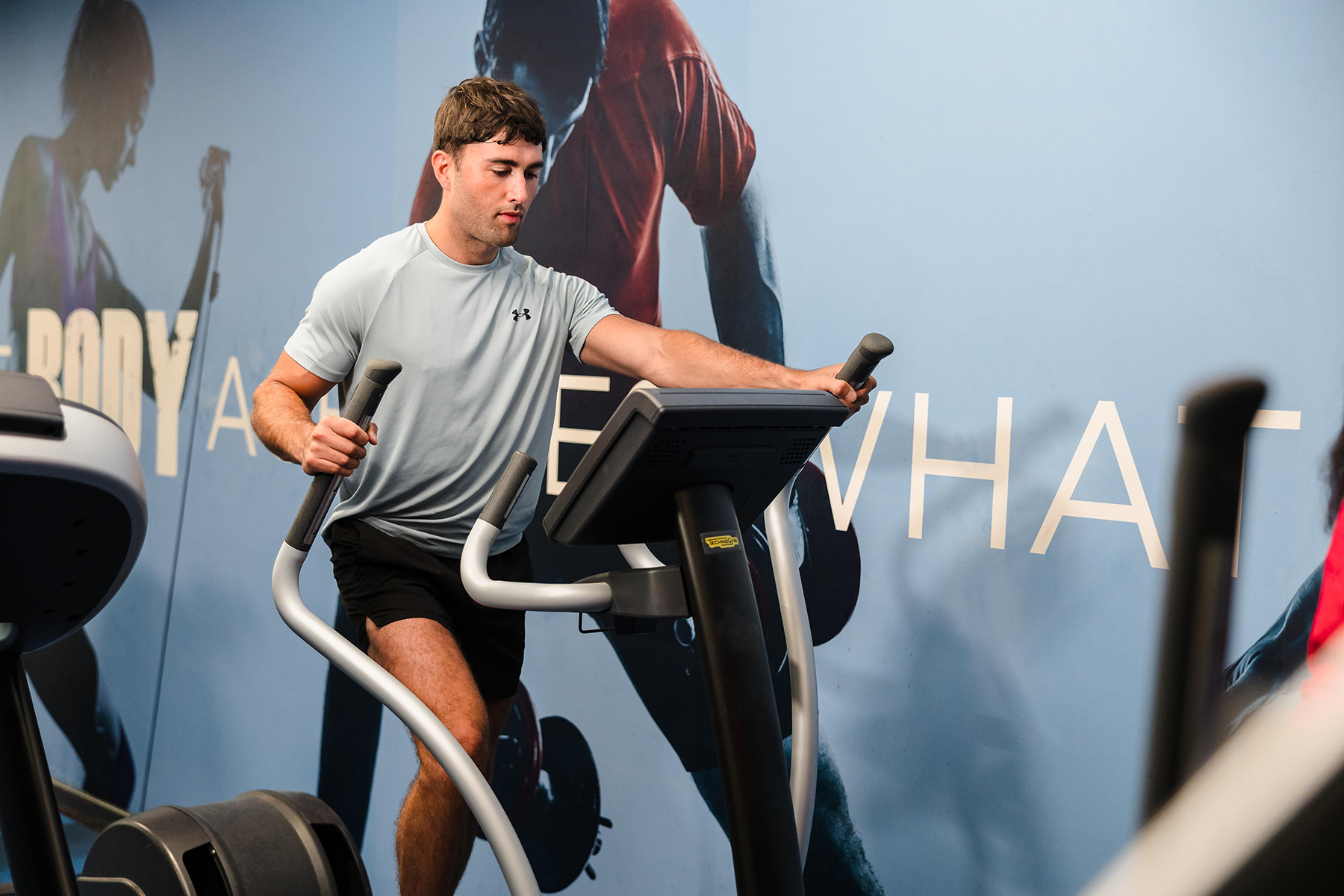 A man is exercising on an elliptical machine in a gym, wearing a light gray t-shirt and black shorts. The background shows a wall with text and blurred images.