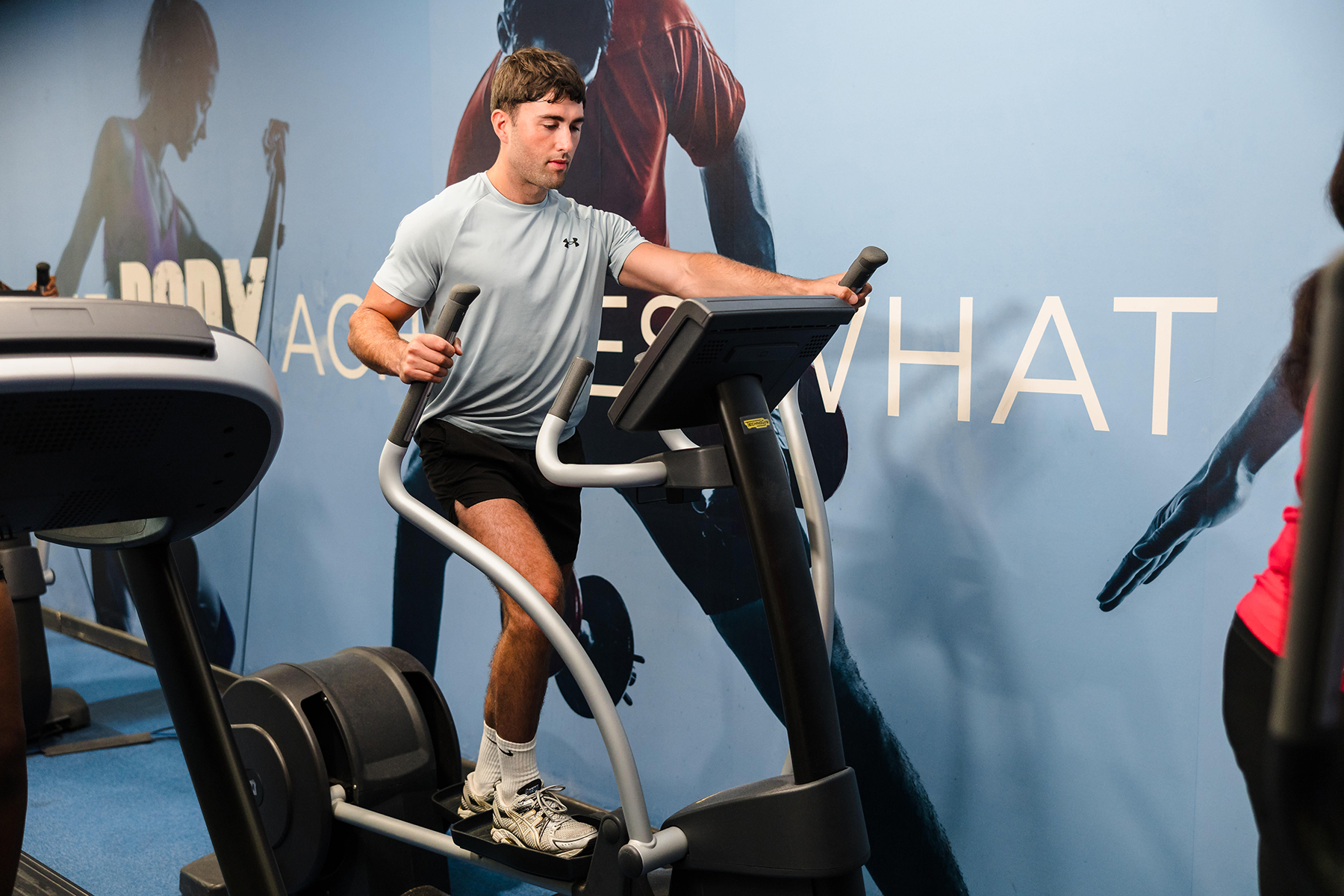 A man in a light gray shirt uses an elliptical machine at the gym, with a motivational wall graphic in the background.