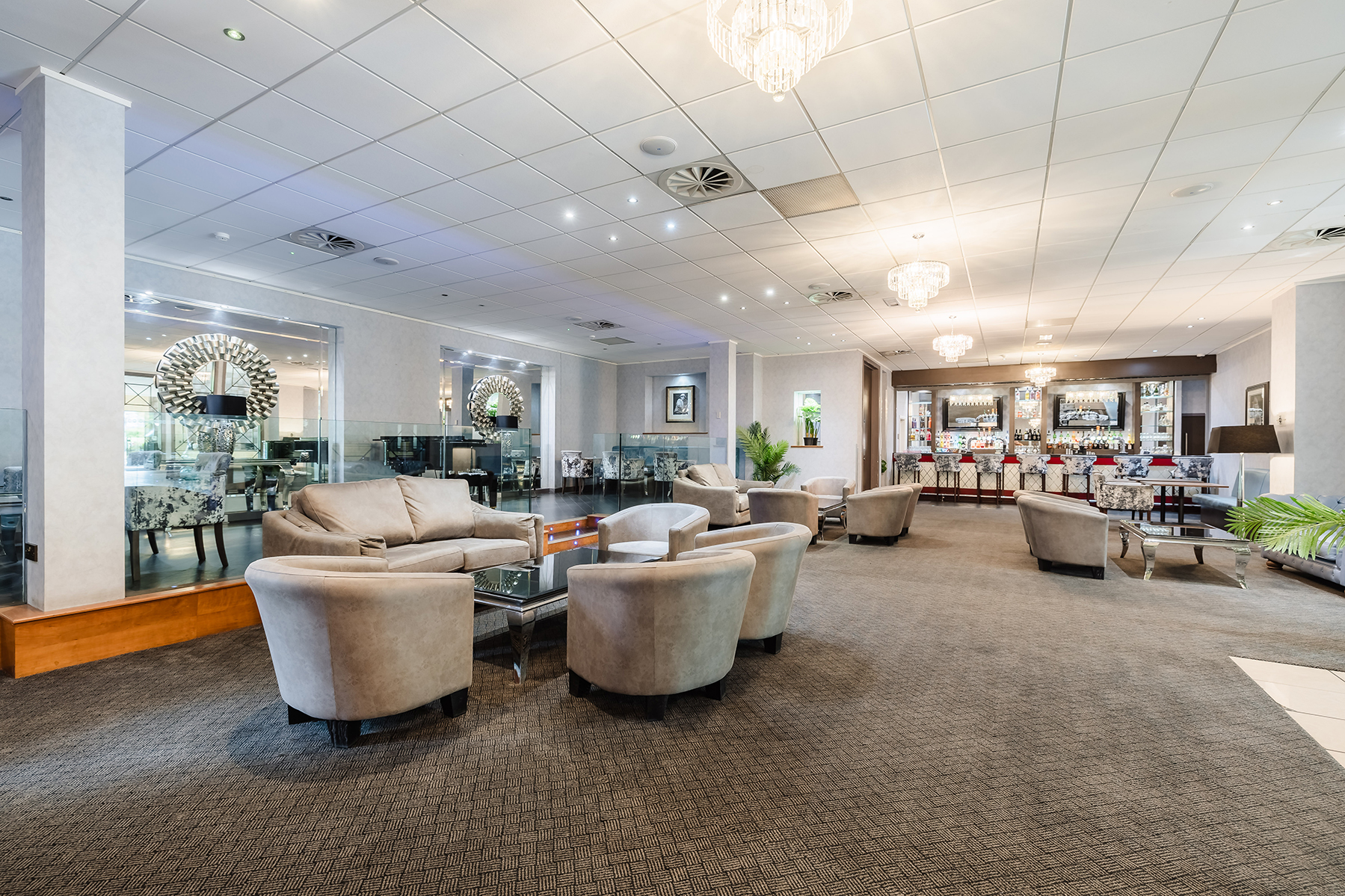 A spacious hotel lobby with beige armchairs, glass tables, decorative mirrors, and chandeliers.