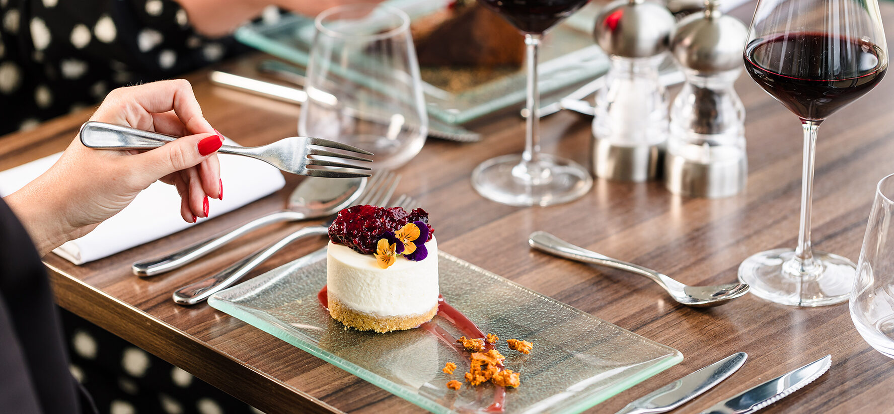 Two people dining at a table with dessert and wine glasses. One person is cutting into a layered dessert with a fork, while the other holds a small white pitcher.