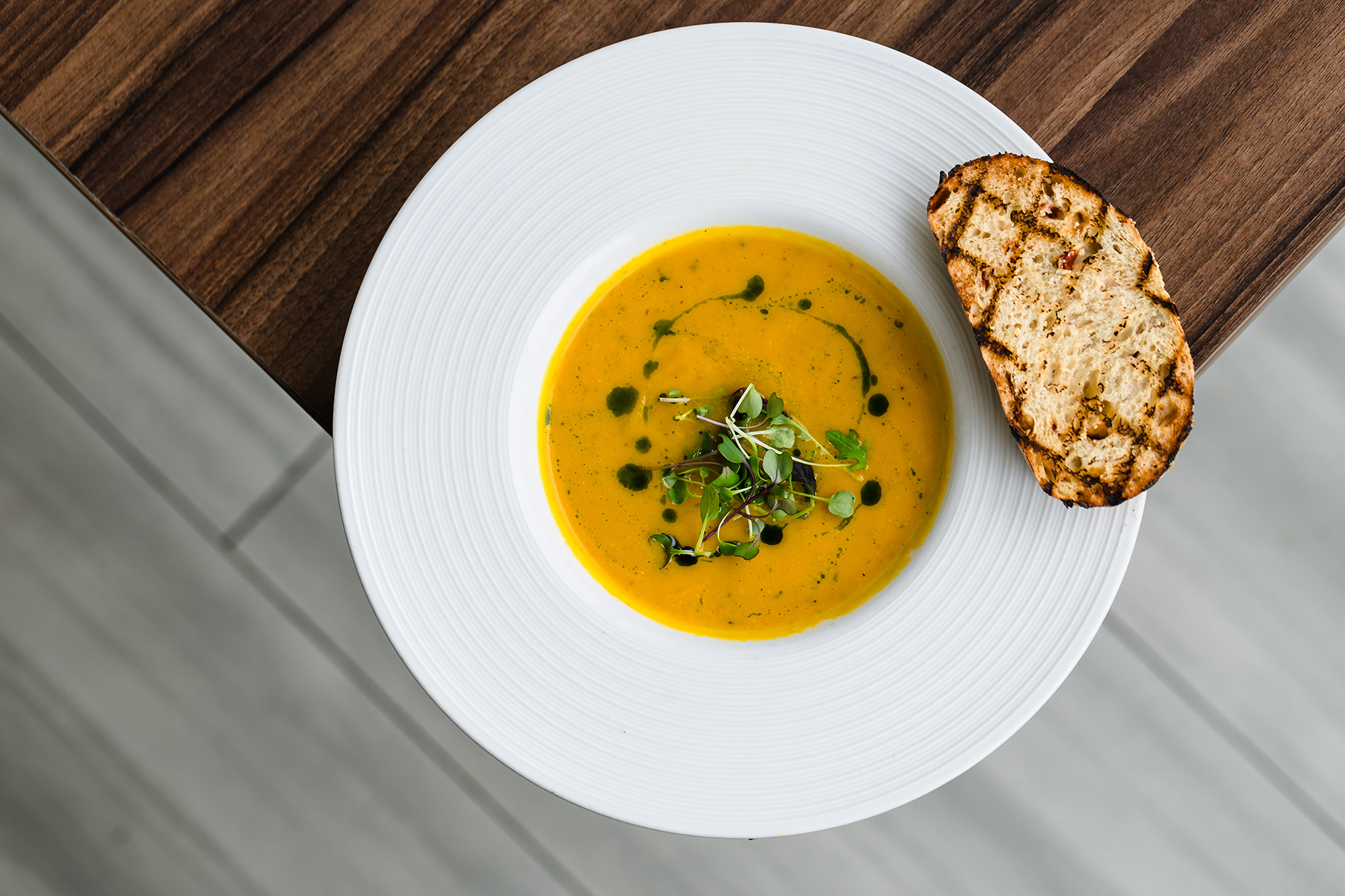 A bowl of orange soup garnished with greens on a white plate, served with a slice of grilled bread on the side.