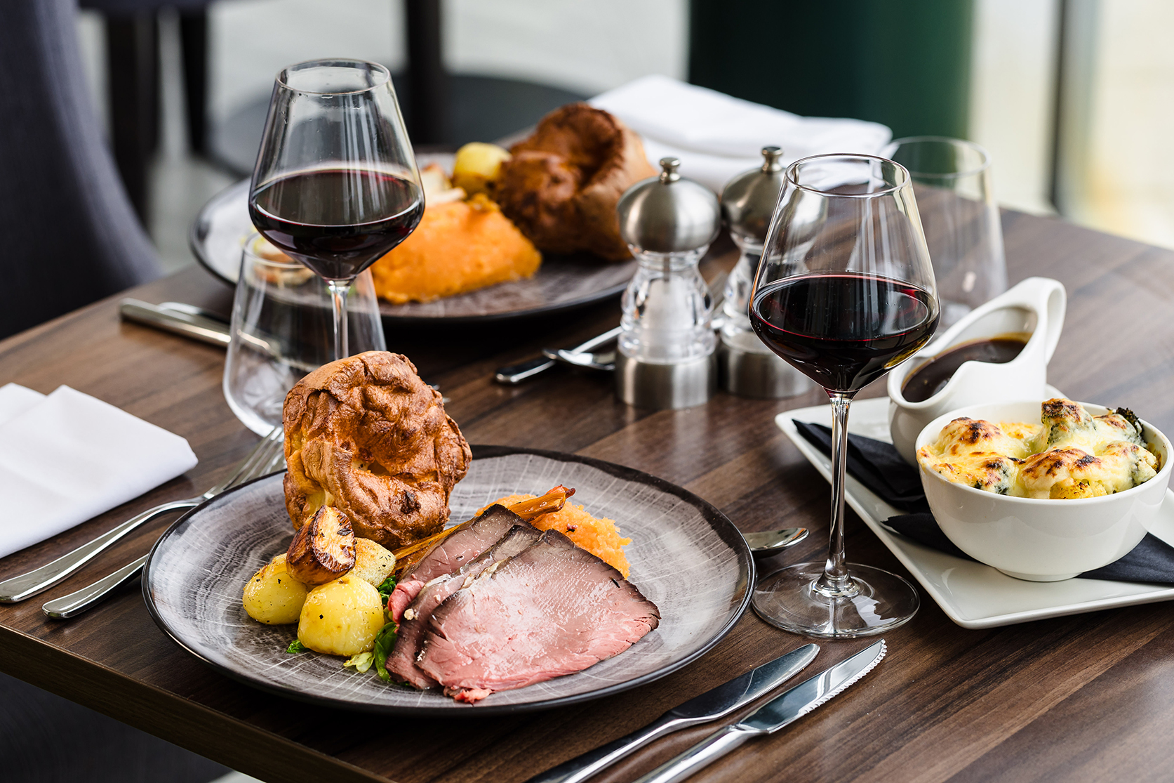 A table setting with two plates of roast beef, potatoes, and Yorkshire pudding, accompanied by glasses of red wine. There is also a dish of vegetables and a gravy boat.