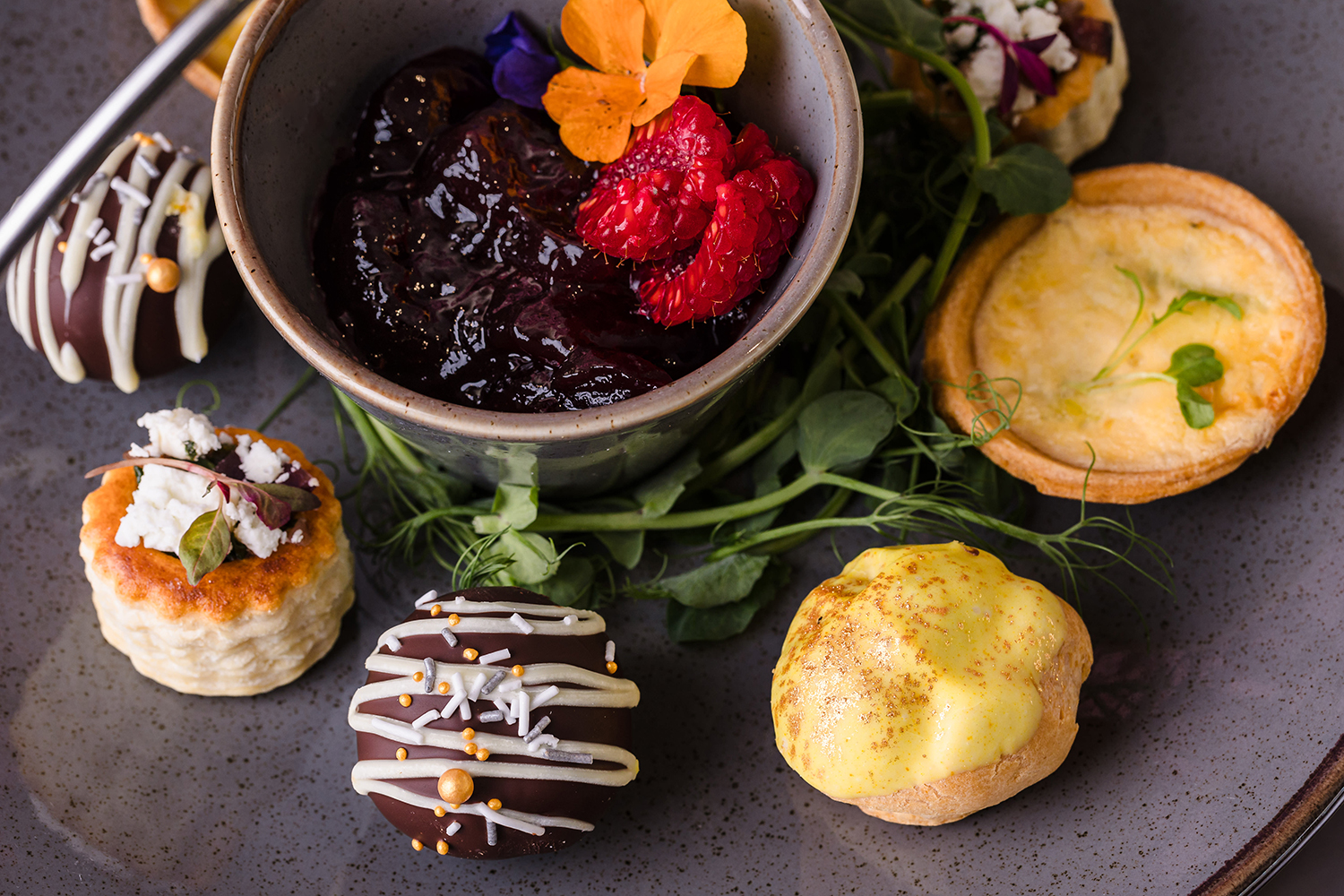 A plate with assorted desserts: a bowl of dark jam with raspberries and flowers, chocolate-covered treats, a mini quiche, and a small pastry topped with yellow cream.