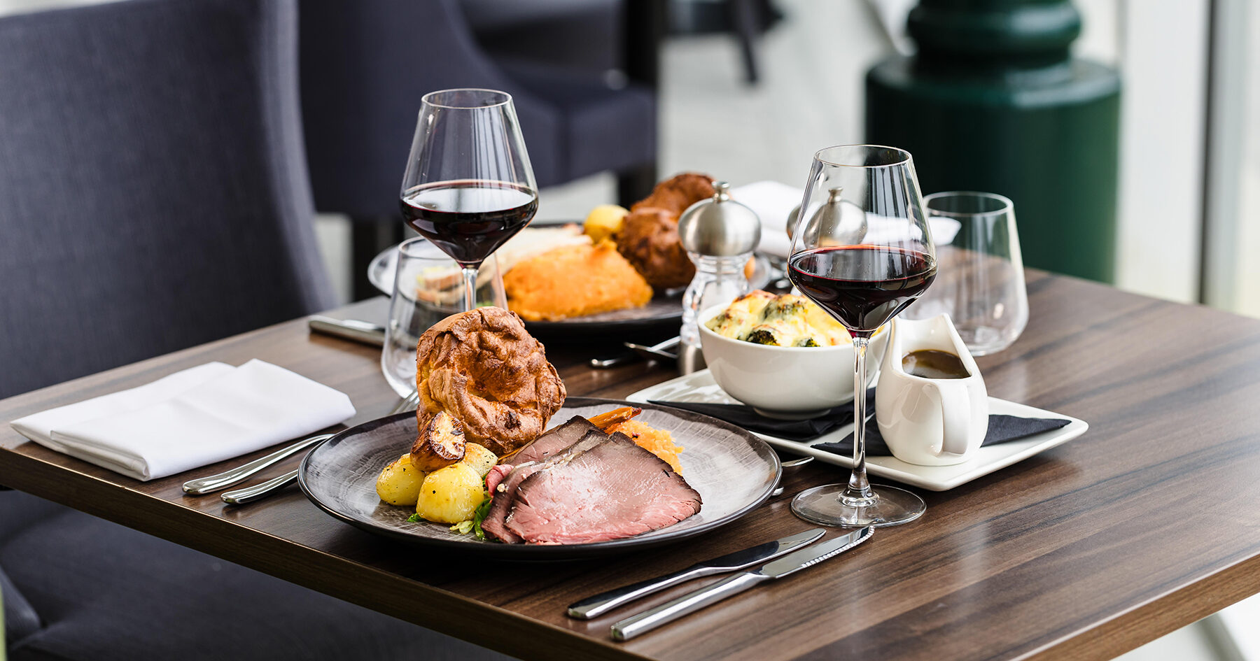 Two plates of roast beef with Yorkshire pudding, potatoes, and vegetables on a dining table, accompanied by two glasses of red wine and a bowl of creamy side dish.