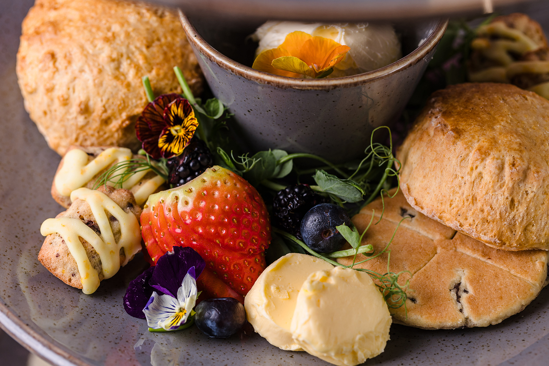 A plate with scones, a strawberry, blueberries, flowers, clotted cream, and a small bowl with a scoop of ice cream.