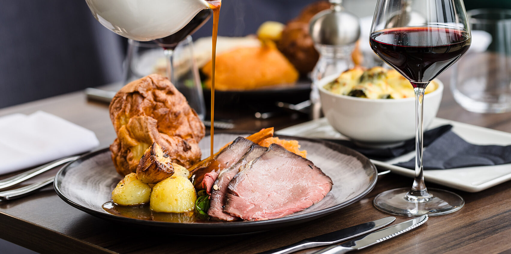 A plate of roast beef with Yorkshire pudding, potatoes, and gravy is served on a table with a glass of red wine.