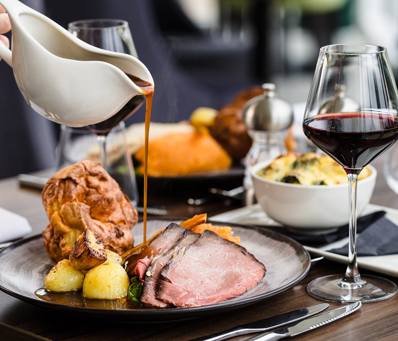 A plate of roast beef, potatoes, and Yorkshire pudding is being drizzled with gravy. A glass of red wine and a bowl of side dish are on the table.