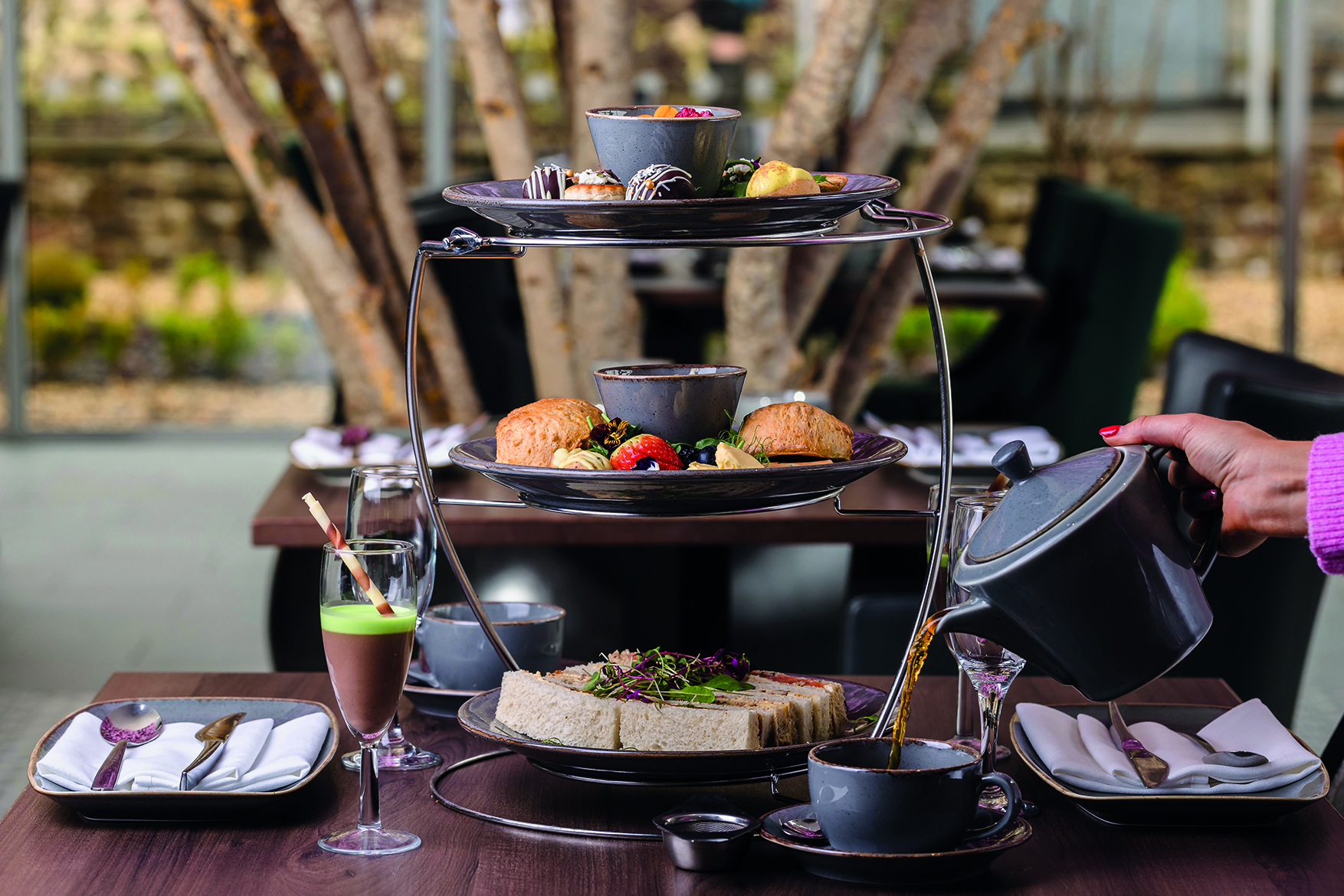 A tiered stand with assorted pastries and scones, a tray of sandwiches below, and a person pouring tea into a cup.