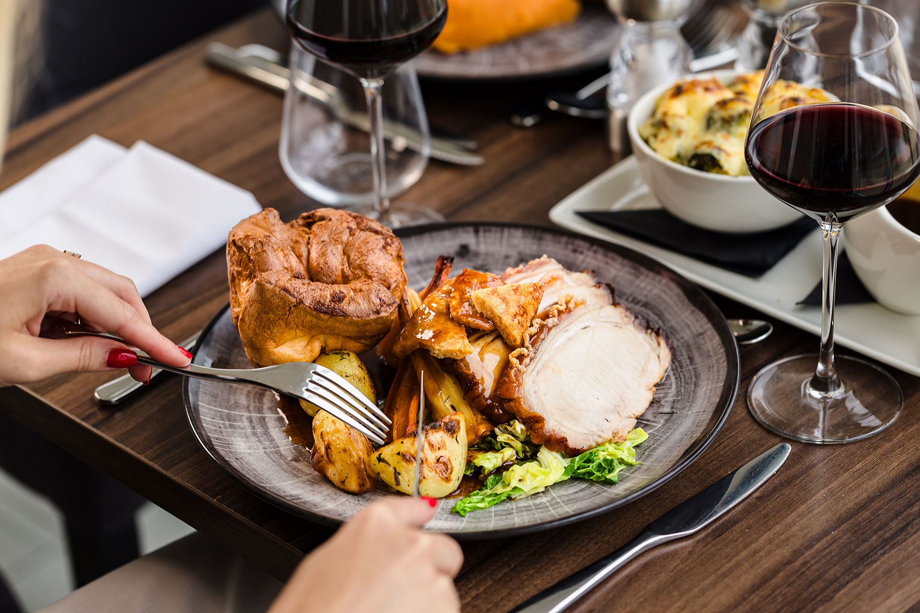 A plate with roast chicken, roasted potatoes, Yorkshire pudding, and greens. A person is using utensils. Two glasses of red wine are on the wooden table.