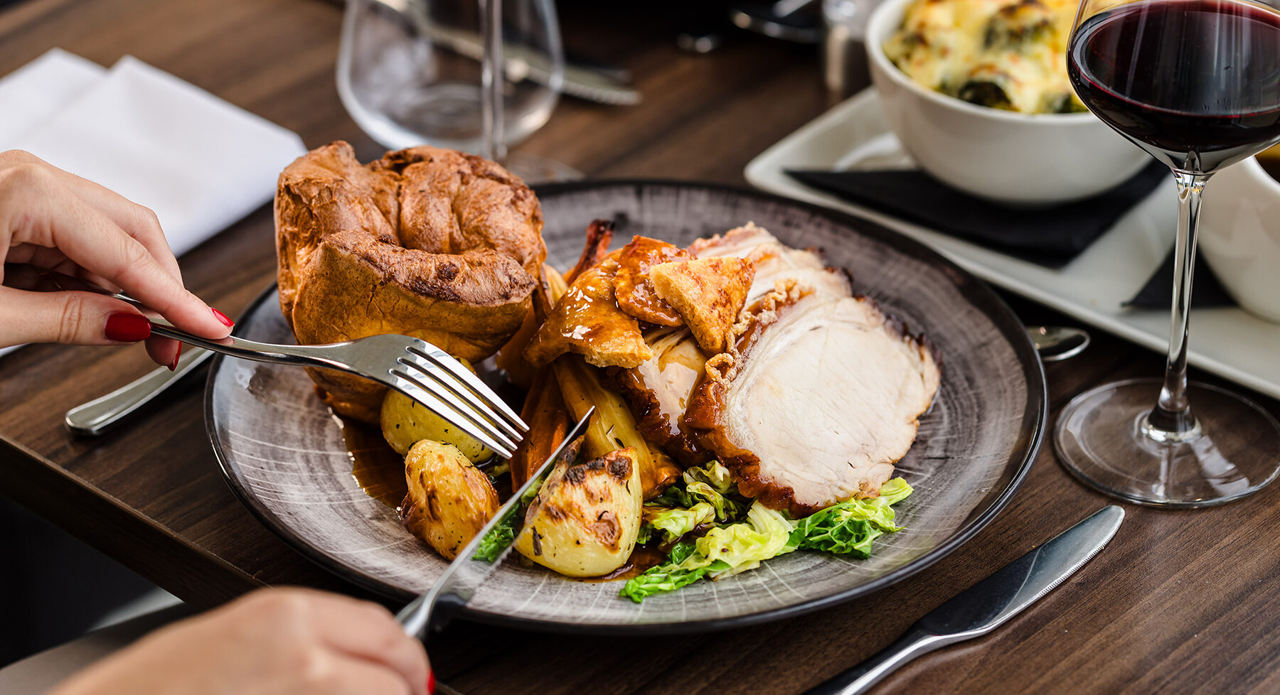 A person is dining on a roast meal with sliced meat, roasted potatoes, cabbage, and Yorkshire pudding. A glass of red wine is on the table.