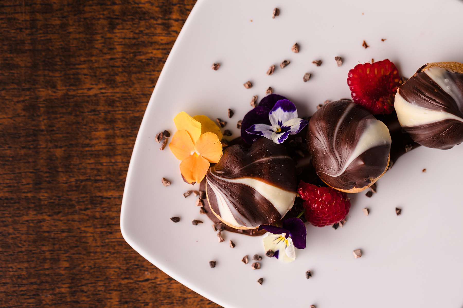 Chocolate-covered pastries with white chocolate swirls, garnished with raspberries, pansies, and edible flowers on a white plate against a wooden background.