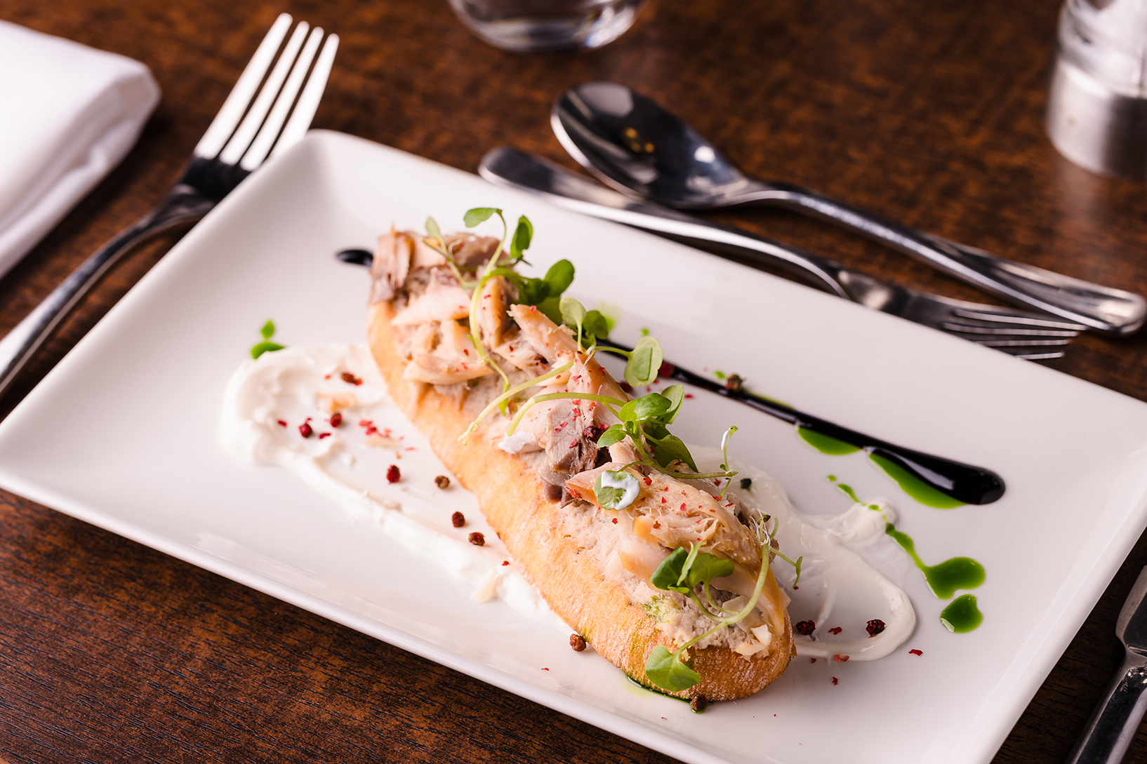 Open-faced sandwich with shrimp, greens, and sauce on a rectangular white plate, set on a wooden table with a fork, knife, and spoon nearby.