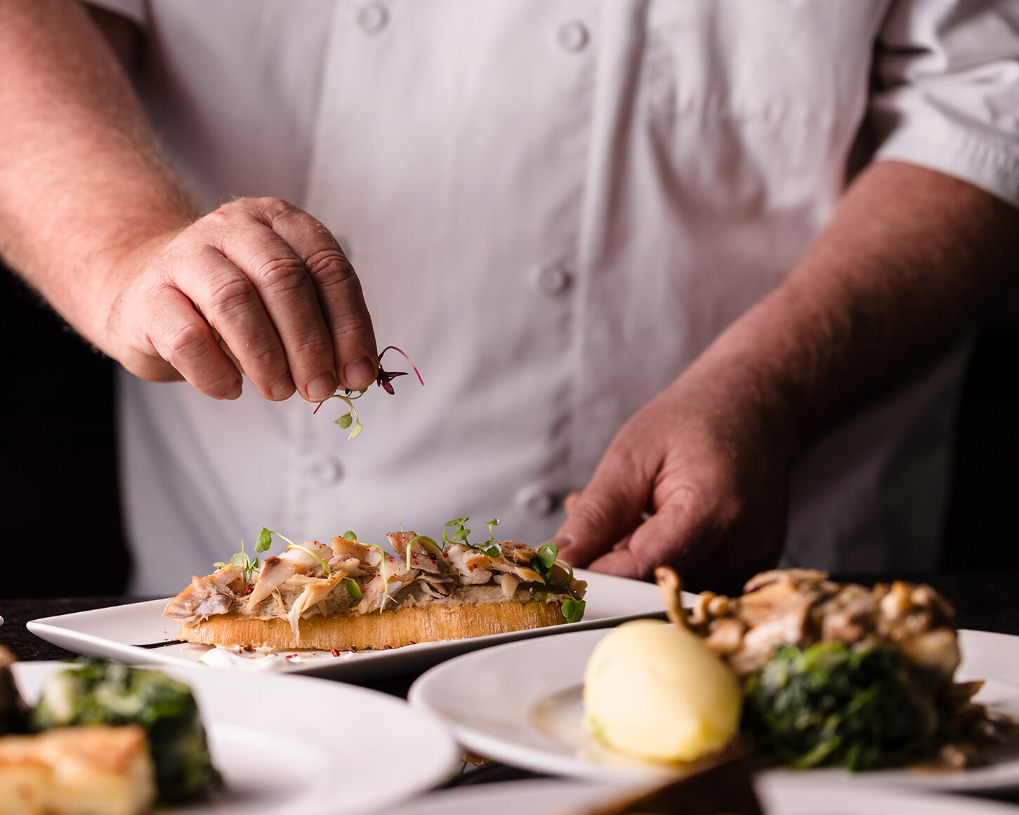 A chef in a white coat garnishes open-faced sandwiches. Plates with dishes, including an egg and greens, are on the table.