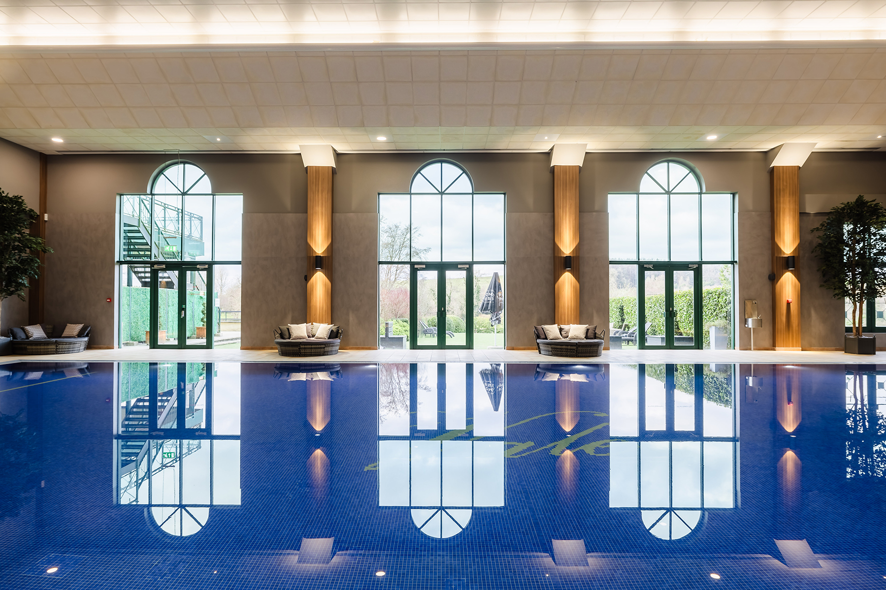 Indoor swimming pool with arched windows, lounge chairs, and plants, reflecting on the calm water surface.