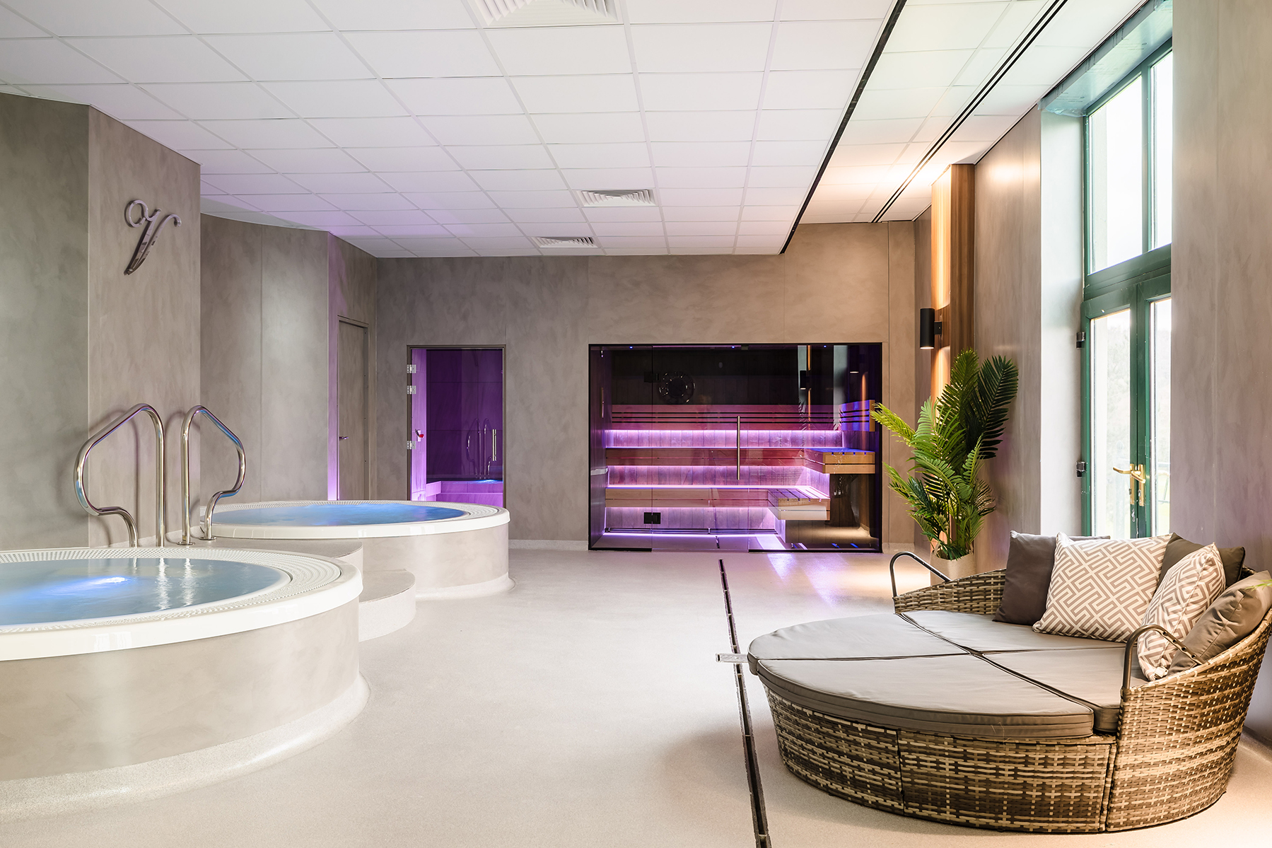 Luxurious spa room with two circular hot tubs, a modern sauna, and a cushioned lounge chair next to a large window.