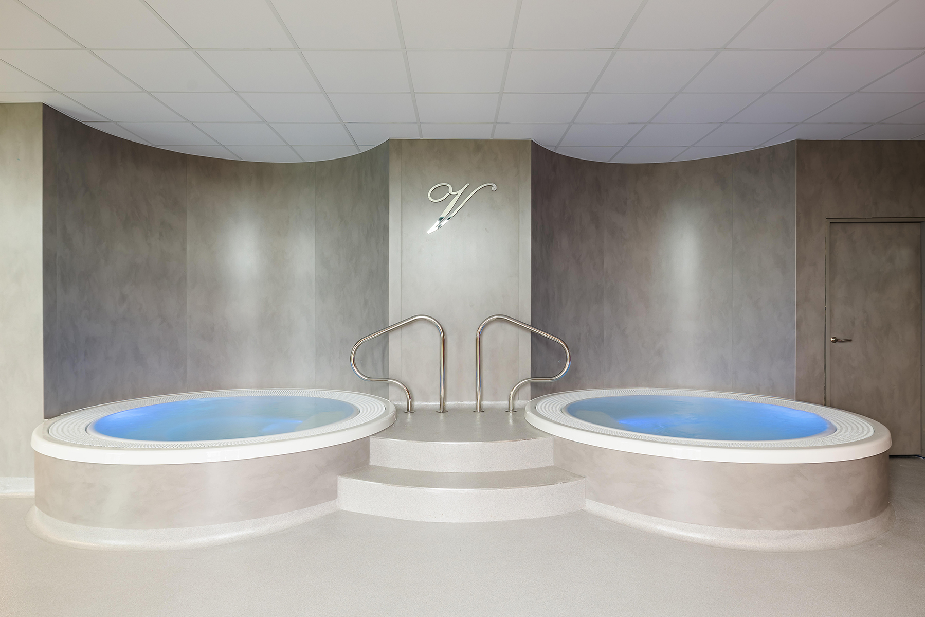 Two indoor hot tubs with blue water, featuring metal handrails and steps, under a ceiling with white tiles.