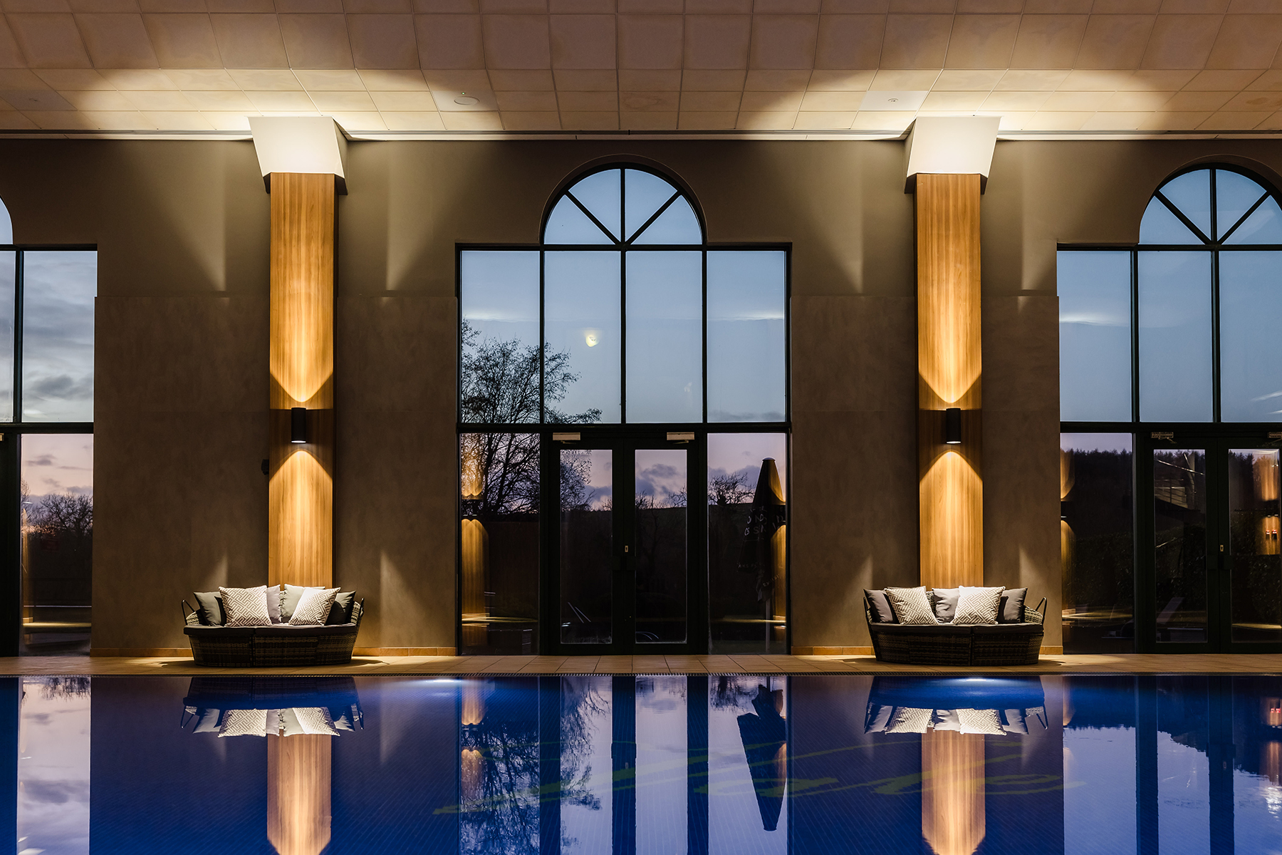 Indoor pool area with large arched windows, cushioned seating, and warm lighting reflecting on the water's surface.