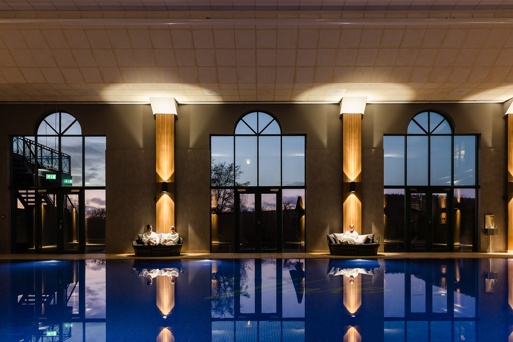 Indoor pool with two people relaxing on lounge chairs by large arched windows reflecting on the water's surface.