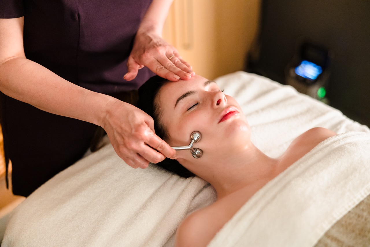 Person receiving a facial massage with a metal roller while lying on a white towel.