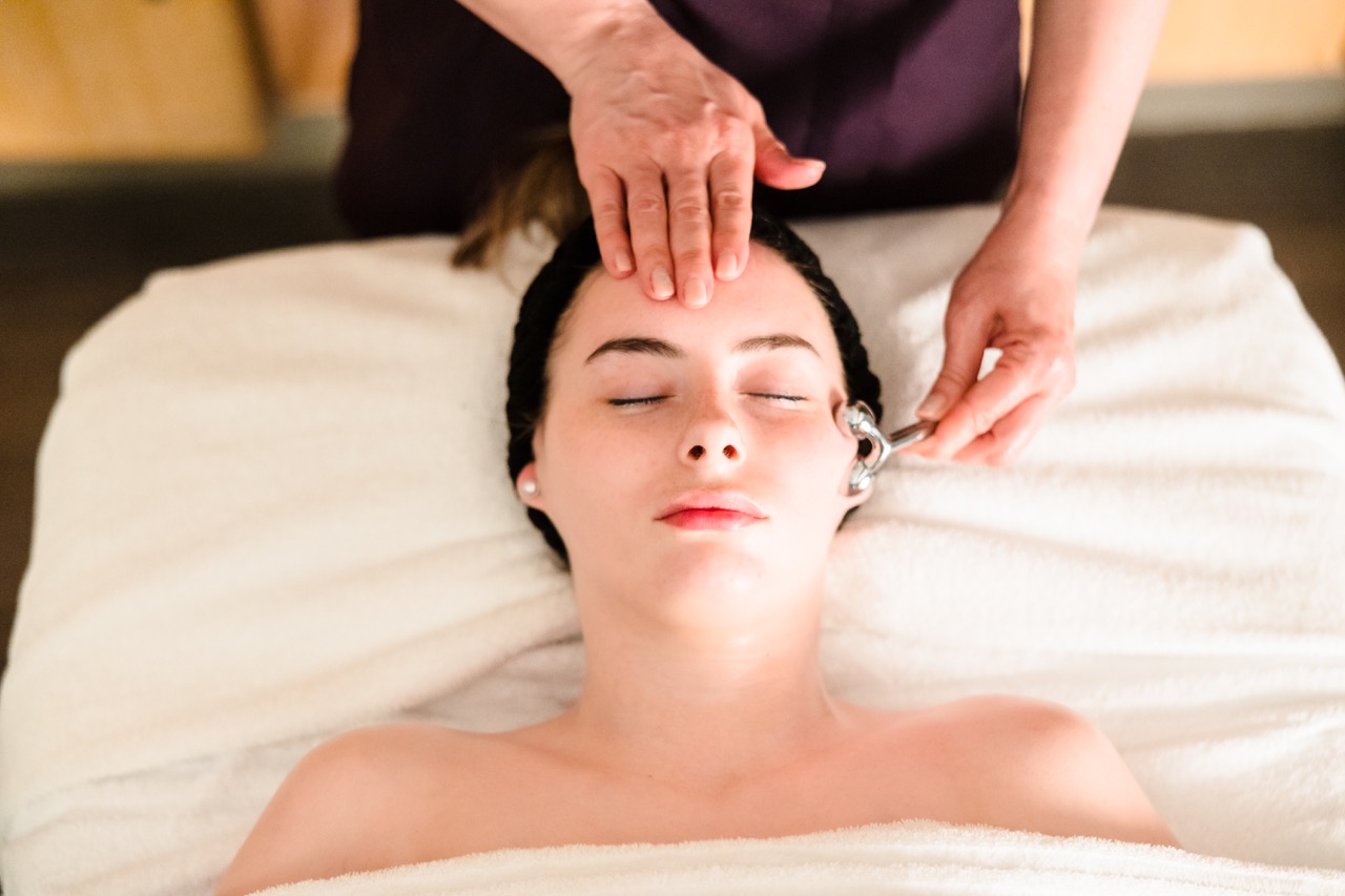 A person receiving a facial massage with a roller, lying on a white towel-covered bed.