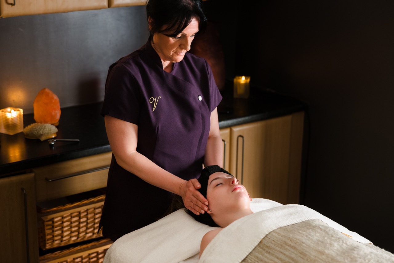 A spa therapist in a purple uniform gives a facial massage to a woman lying on a massage table in a dimly lit room with candles.